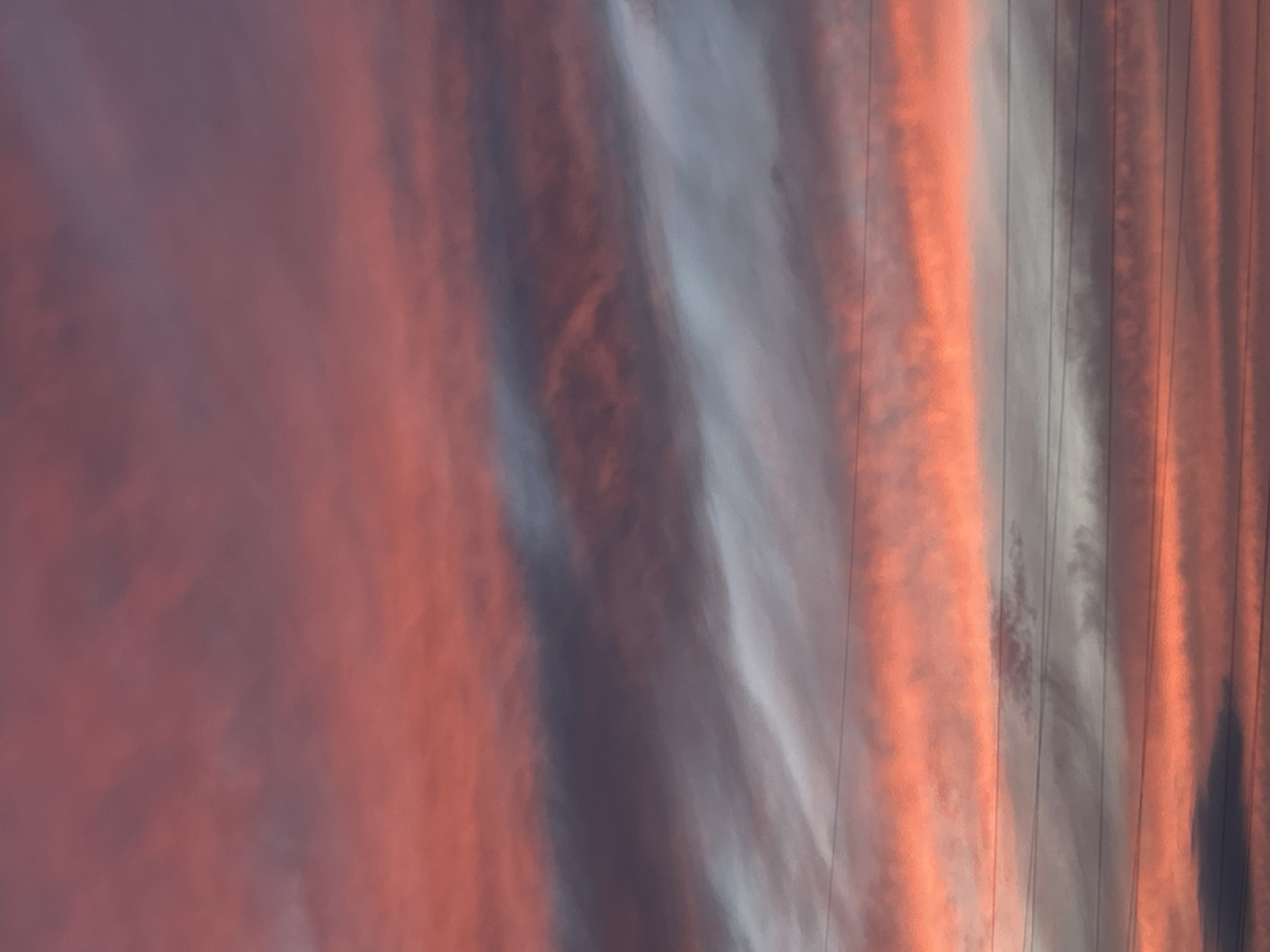 The image is an outdoor shot of a sky filled with striated clouds during what appears to be sunset or sunrise. The clouds exhibit a range of colors from deep reds and oranges to grays and whites. Several thin, horizontal lines, likely power lines or cables, are visible across the frame, interrupting the cloud formations. There are no people or identifiable landmarks in the image. The scene depicts a natural atmospheric phenomenon without any specific urban context visible.