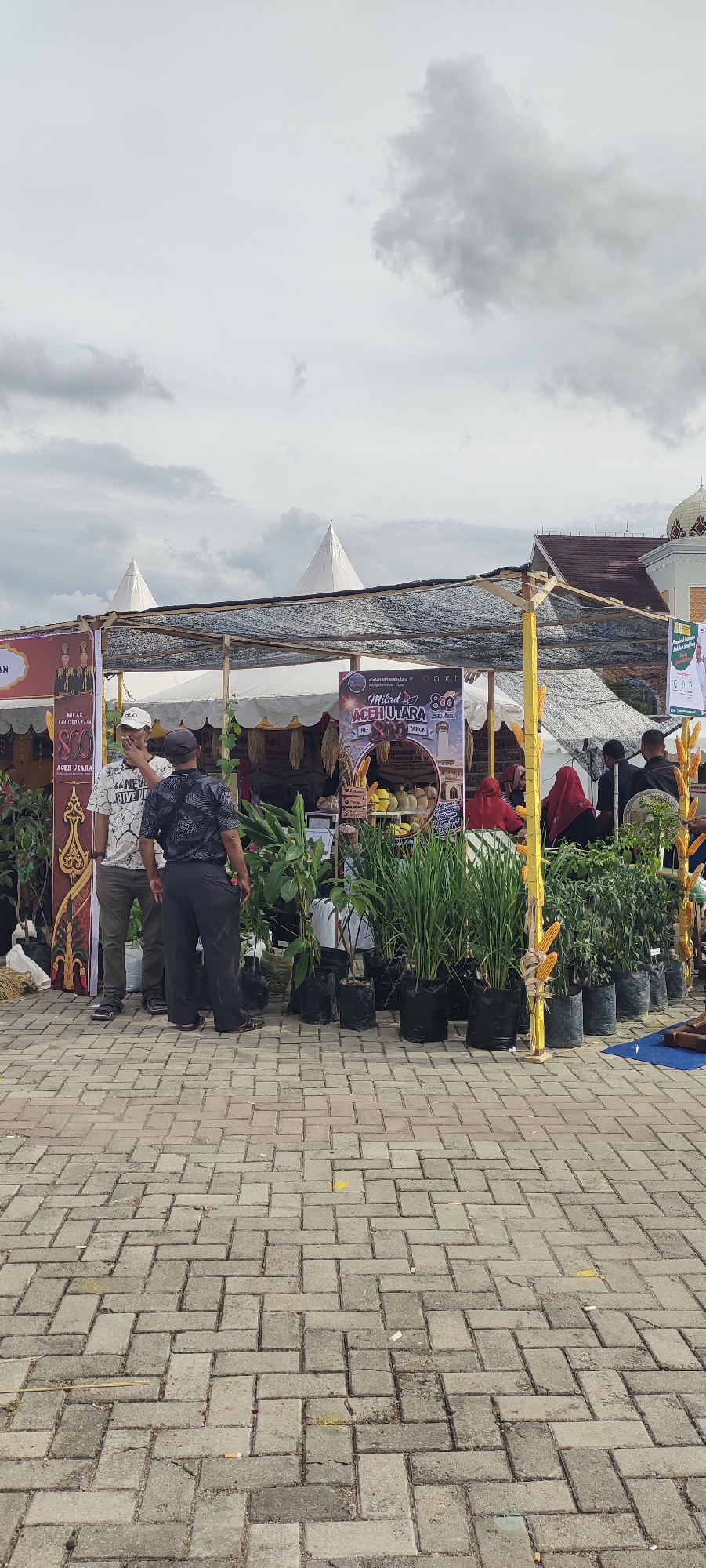 The image depicts an outdoor market stall in Meunasahaluedrienlhoksukon Barat, Indonesia. The stall is covered by a dark mesh tarp supported by yellow poles. In front of the stall, several people are visible. Two men in the foreground are looking towards the stall. One is wearing a white t-shirt with a graphic print and olive-green pants, while the other is in a dark, patterned short-sleeved shirt and dark pants. Further back, several individuals, some wearing headscarves, are interacting at the stall.
The stall itself is filled with various plants in black bags, including what appears to be rice stalks, leafy green plants, and some flowering plants. There are also some fruits, possibly durian and melons, displayed on a table. Banners with text and images are hung at the stall, one of which features a circular graphic and text in what appears to be Indonesian. In the background, two white conical tents are visible, as well as a building with a dome and ornate architectural details, suggesting a religious or governmental structure. The ground is paved with interlocking bricks. The sky is overcast with scattered clouds.