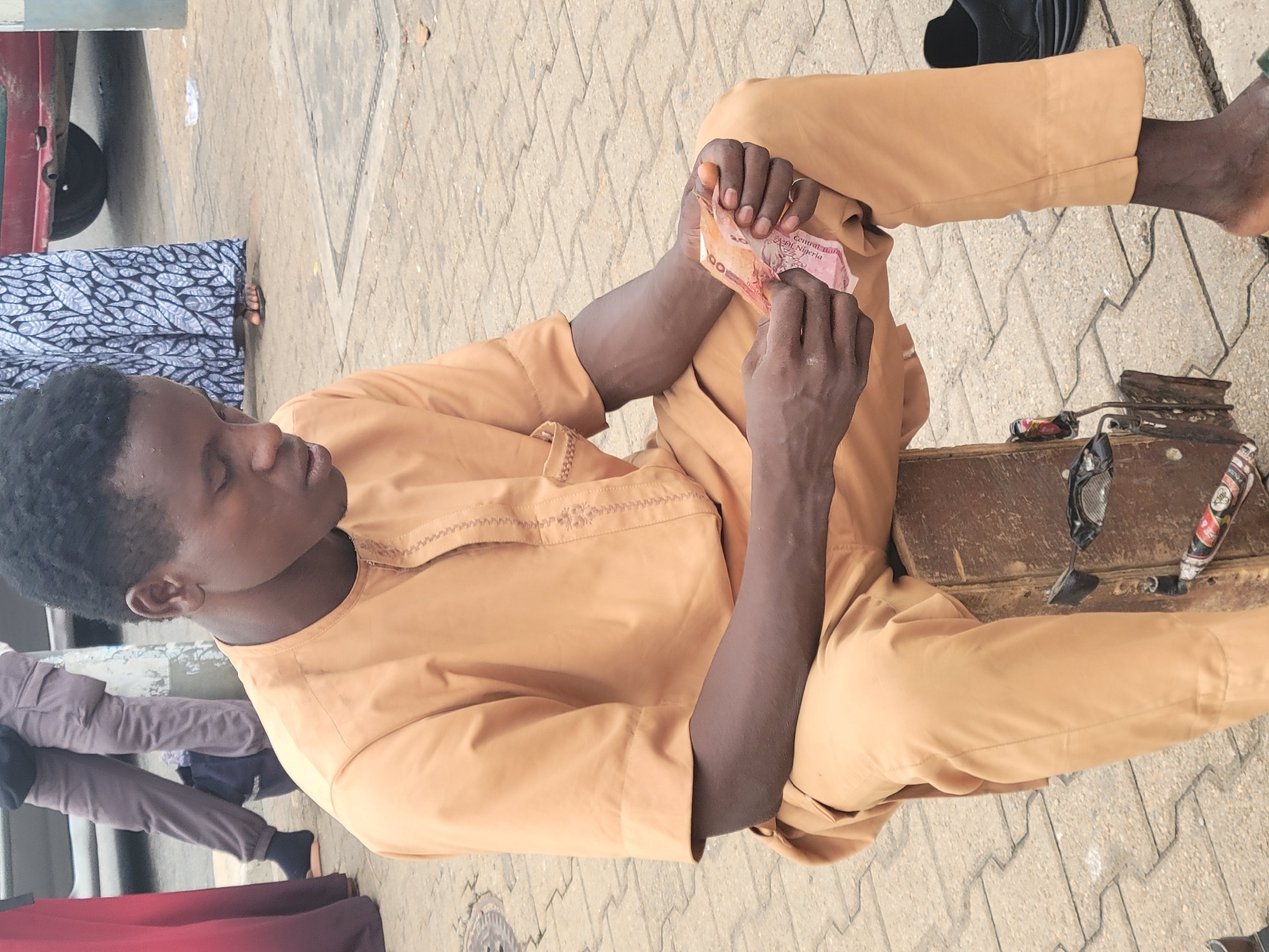 A man in a tan outfit sits on a brown bench and examines a 100 naira note. He has short, dark hair and is dark-skinned. The bench is located on a paved area with a geometric pattern. To the right of the man is a small toolbox and a pair of sunglasses. Other people are visible around the man. The location is likely Abuja, Nigeria, based on the currency.