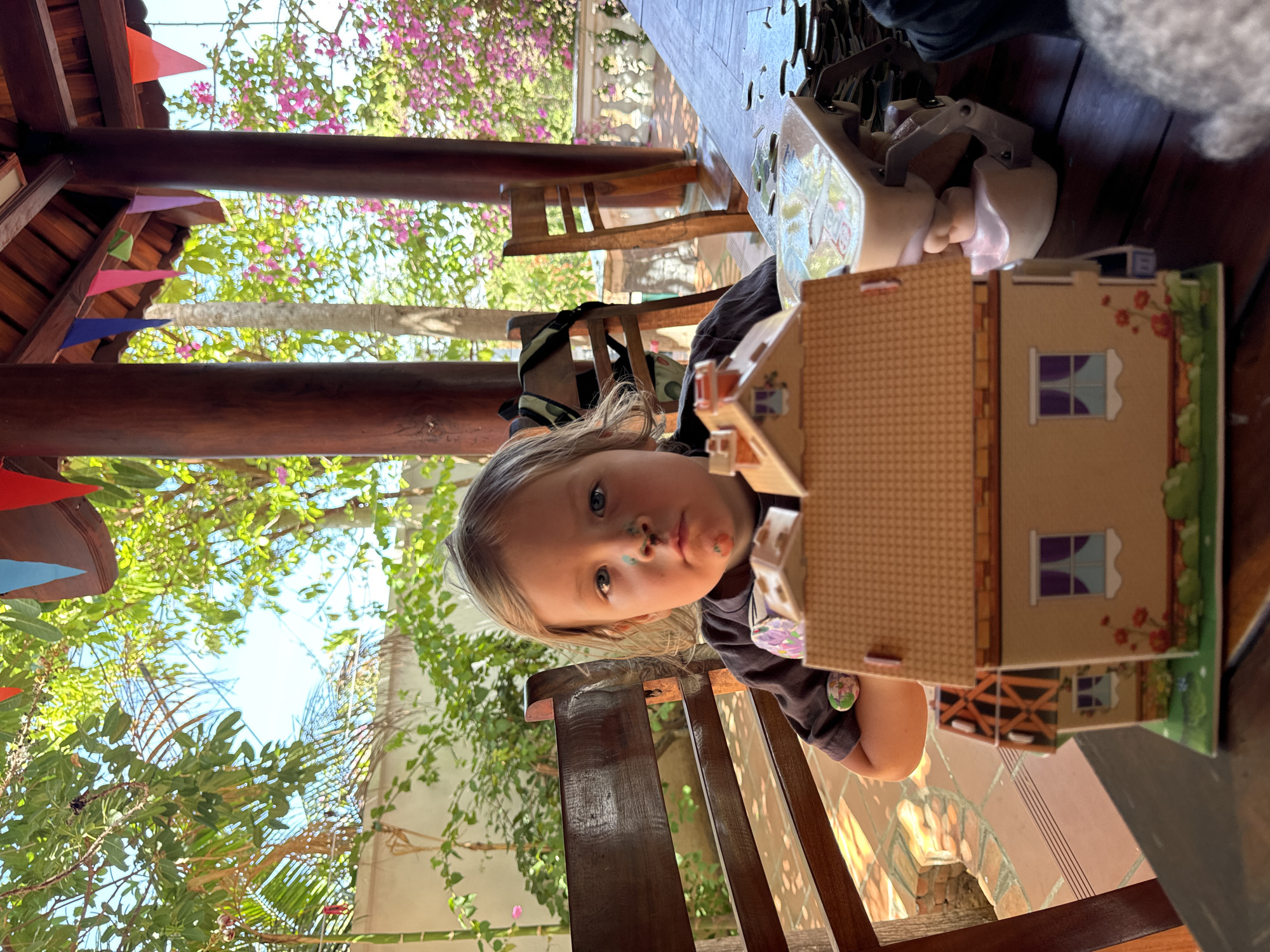 A low-angle shot captures a young child with paint smudges on their face seated at a wooden table and chair in Mũi Né, Vietnam. The child, with blonde hair, is holding a cardboard house in front of them. The setting appears to be a patio or outdoor seating area, with vibrant greenery and flowering vines visible. Colorful pennant flags hang from the wooden structure above. A portion of a wooden table is visible, with items like a purse and other objects placed on it. The flooring is patterned tile. The lighting suggests daytime, with a mix of sunlight and shadow.