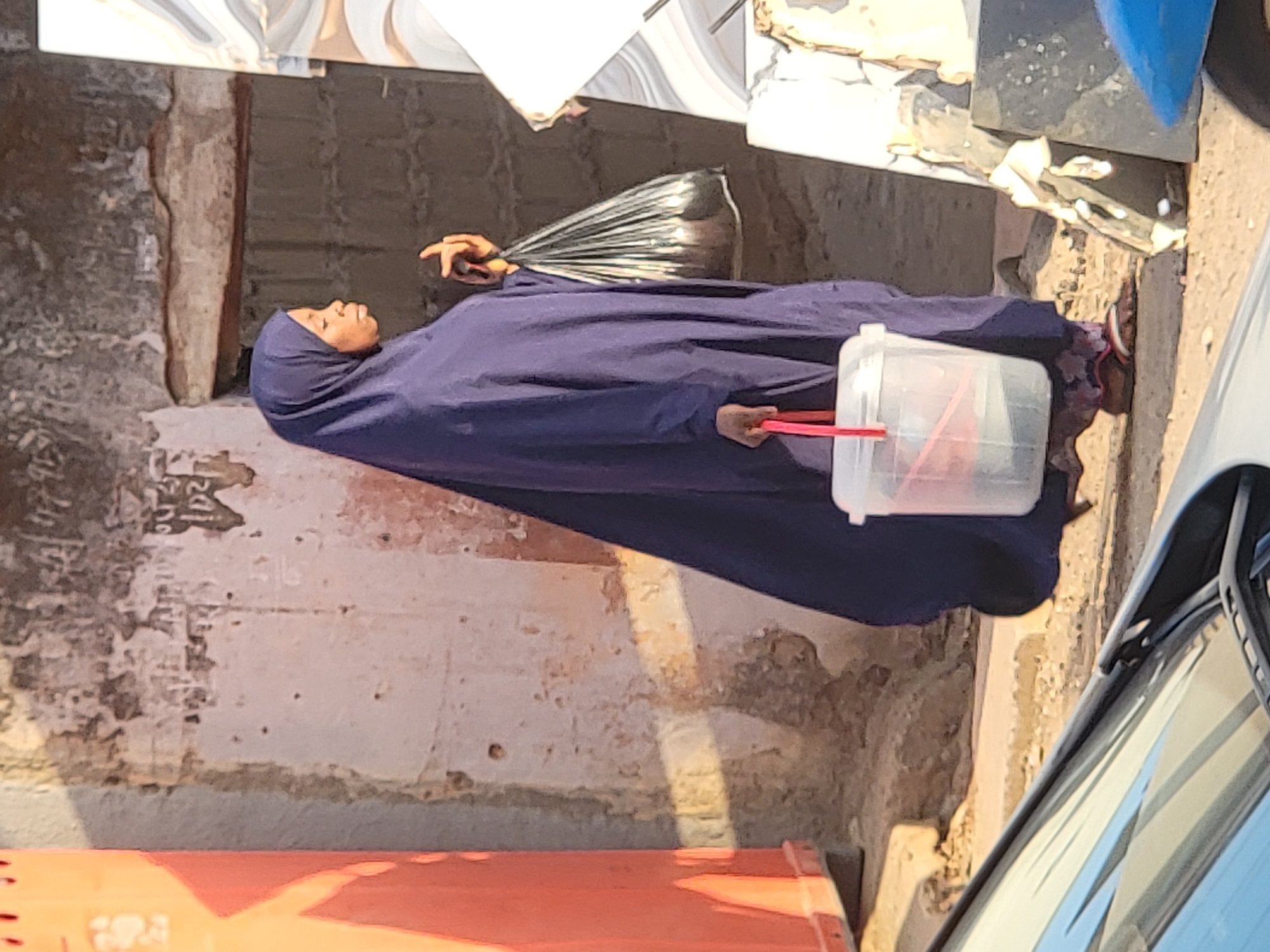 A woman in Maiburiji, Nigeria, dressed in a blue hijab and flowing garment, lies on a concrete surface near a crumbling wall. She is holding a plastic bag in one hand and a translucent plastic container with a red handle in the other. Part of an orange container is visible near the bottom left corner and a portion of a vehicle is on the right. Tiled surfaces are stacked above her.