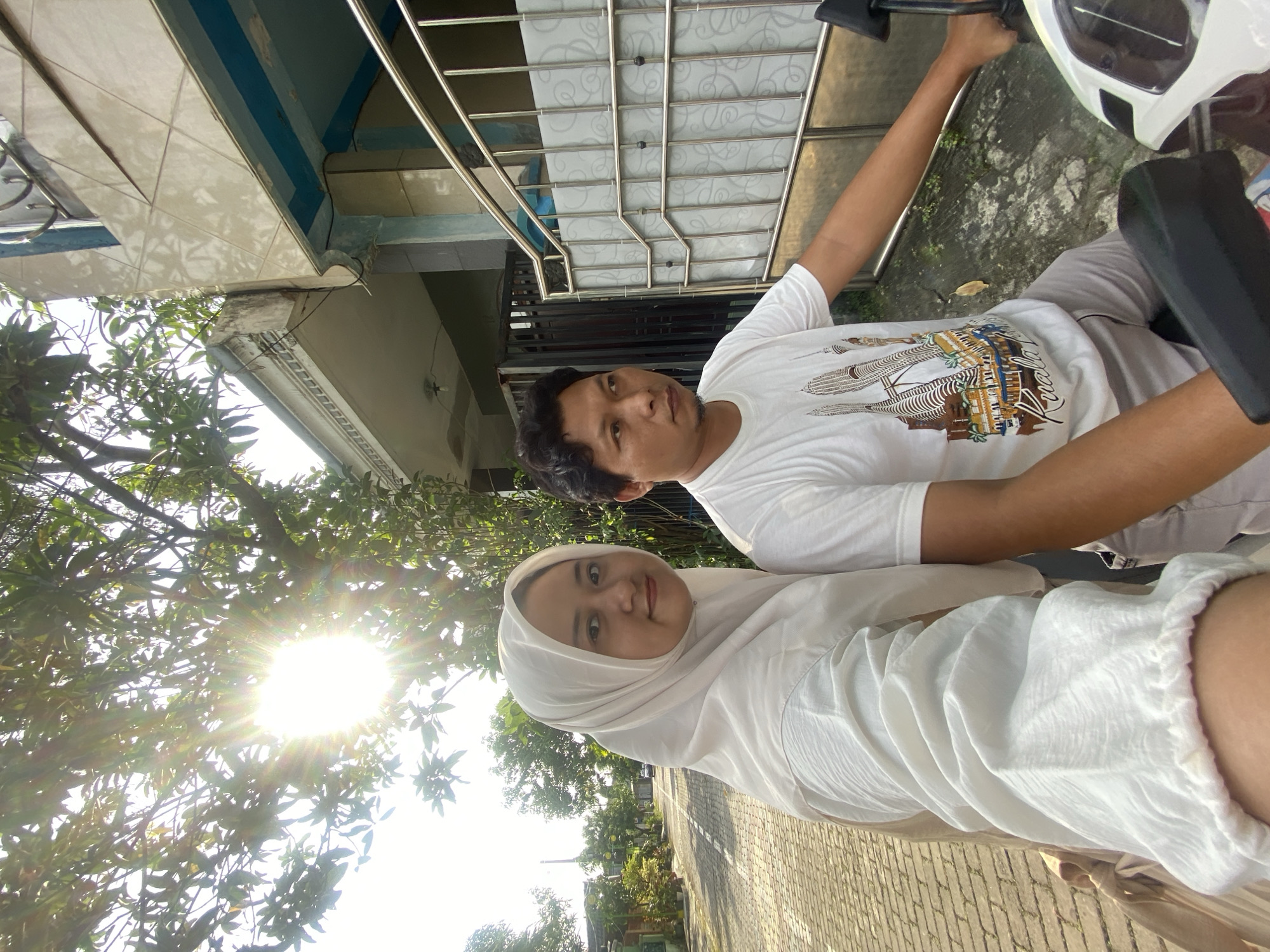 A man and a woman are on a parked moped on a brick sidewalk in Gelam, Indonesia. The man wears a white t-shirt with an illustration of the Petronas Towers on it, and grey pants. The woman wears a white hijab and white clothing. The sun shines through trees in the background. A building is partially visible in the upper left. A metal canopy is overhead.