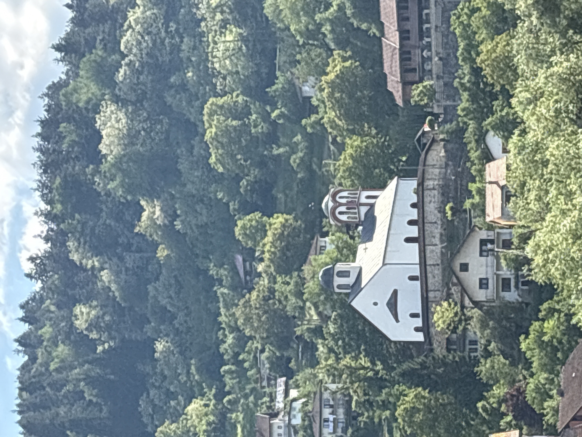 The image shows a white church with domes, surrounded by lush green trees and vegetation. The church is located on a hillside, with residential buildings visible in the background. A dense forest covers the landscape behind the church. The sky is partially visible in the upper left corner, with some clouds present. The orientation of the photo suggests it was taken from an elevated position. Without further context, the specific location within Foča, Bosnia and Herzegovina cannot be definitively determined.