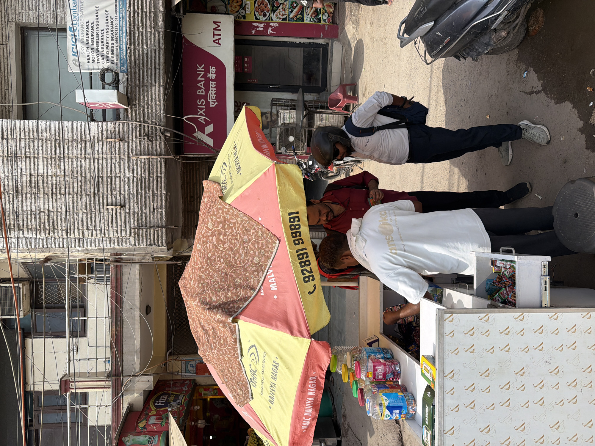 The image depicts a street scene in Malviya Nagar, Delhi, India, on a sunny day. In the foreground, a vendor's stall is set up under a large, colorful umbrella that is red and yellow. The umbrella is patterned with a brown, floral design on its upper section. The stall itself is a white counter with a drawer, displaying various products like bottled drinks and packets of snacks.
Three men are interacting at the stall. One man, wearing a white t-shirt, is behind the counter, appearing to serve a customer. Another man, in a red shirt, stands to his left, looking towards the counter. A third man, wearing a white shirt, dark pants, a helmet, and sunglasses, is facing the counter from the right. He has a backpack on.
Behind the stall, there is a building with a facade featuring a prominent "AXIS BANK" sign with an ATM machine. Further up the building, there is another sign for "ICICI Lombard General Insurance." The architecture suggests a commercial and residential area, with balconies and numerous electrical wires crisscrossing the scene. To the left of the stall, a portion of another shop, "Fiza Juice Shakes," is visible. A motorcycle is parked to the right of the frame. The ground is paved and appears to be a street or sidewalk.