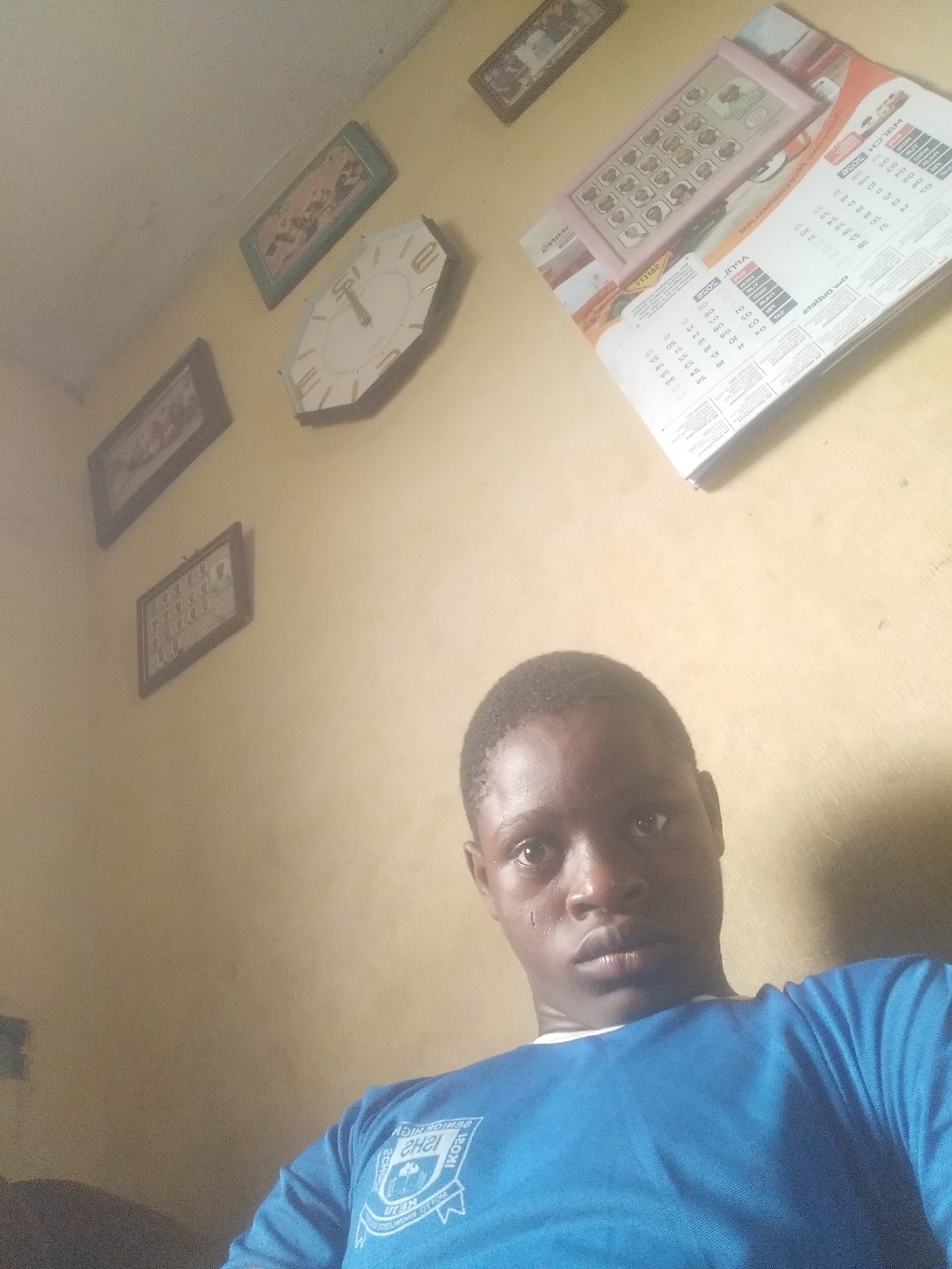 The image presents a low-angle, self-portrait of a young Black man wearing a blue t-shirt with a white logo. The logo on the shirt appears to have the text "SENIOR HIGH SCHOOL". The wall behind him is painted a yellowish color and features several framed pictures, a non-circular clock, and some stacked paper materials, potentially calendars or pamphlets. The location, if in Shangisha, Nigeria, cannot be verified from the image content alone. The scene is indoors.