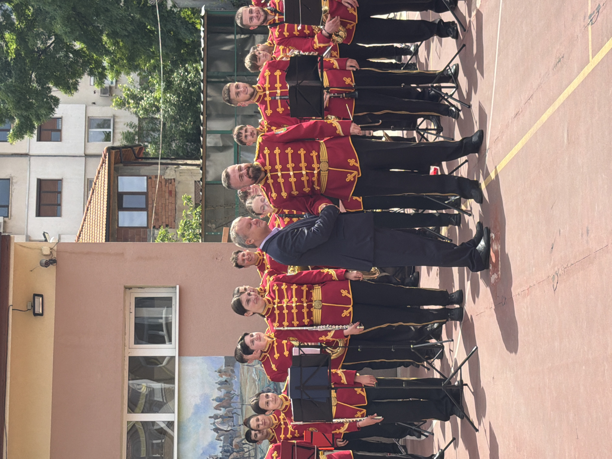 A color photograph shows a brass band standing in formation outdoors, presumably in Pleven, Bulgaria. The band is arranged in a line on a pinkish-red surface, likely asphalt, marked with yellow lines. A man in a dark suit stands amidst the band members. The band members wear red and gold ornate jackets, black pants, and black shoes, with music stands in front of them. Some hold flutes.
In the background, there's a wall with a painted mural and a window. Further back are buildings with trees obscuring parts of them.