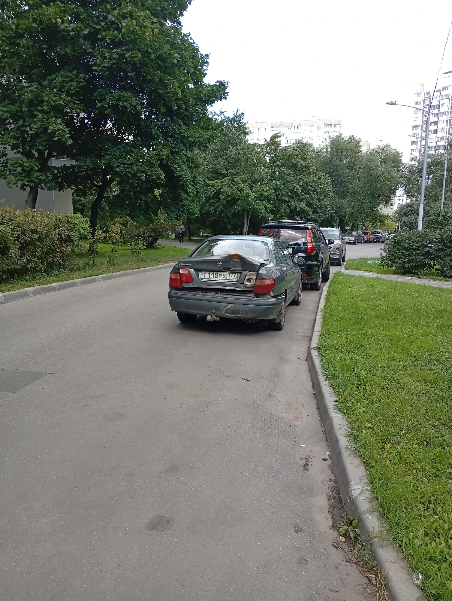 A line of parked cars is visible on the right side of the road. The closest car, a dark gray sedan, has significant damage to its rear bumper and trunk. Its license plate reads "T 118PX 177". Behind it, other vehicles, including an SUV and several sedans, are parked along the curb. The road is paved and appears to be a street or residential area. To the right of the parked cars, a strip of well-maintained green grass runs along the sidewalk, bordered by a concrete curb. Large, leafy trees line the left side of the road, casting shadows. In the background, modern apartment buildings are visible. The sky is overcast.