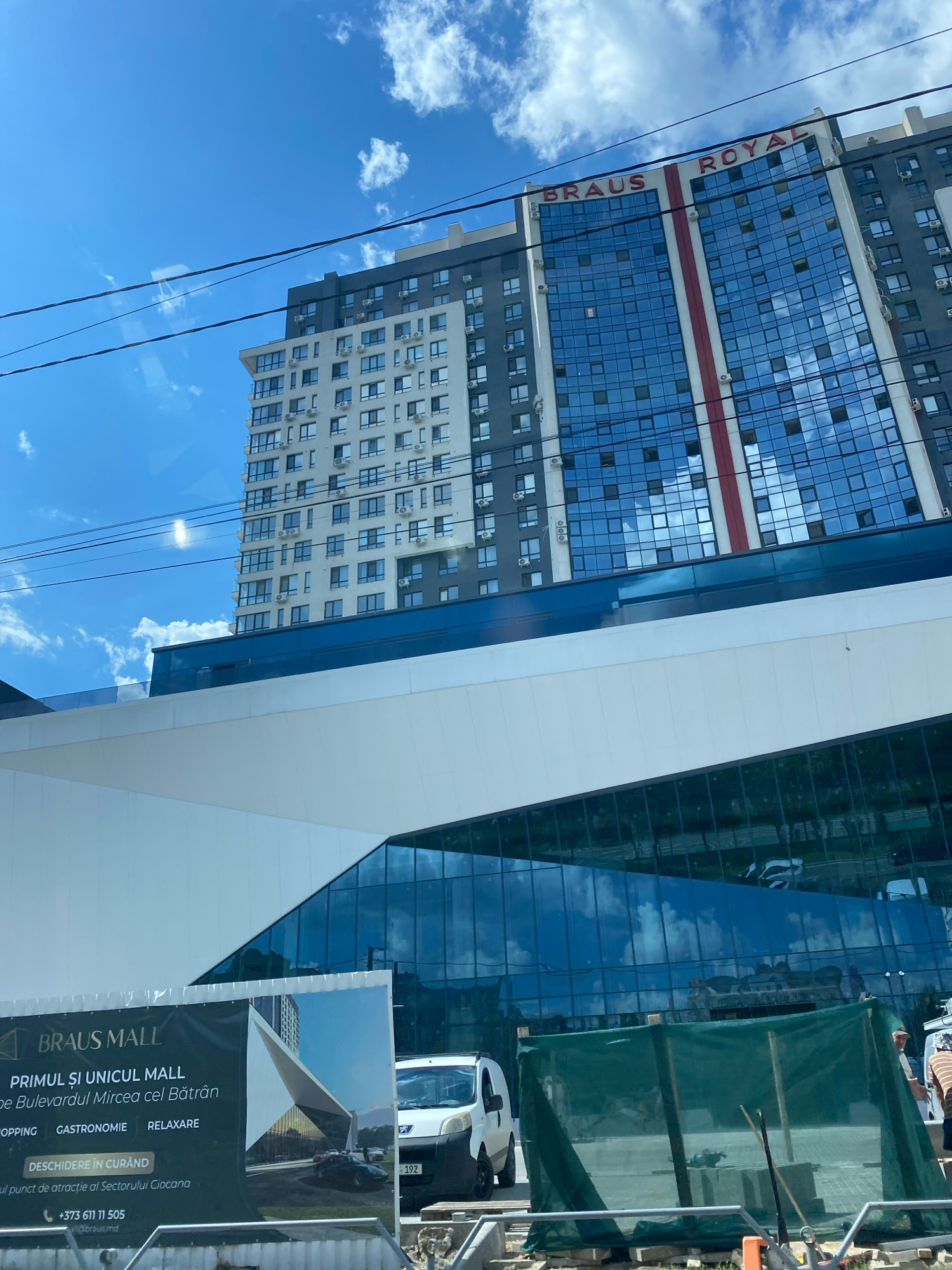 The image shows the Braus Mall under construction in Chișinău, Moldova. The mall is a modern building with a large glass facade that reflects the blue sky and white clouds. The Braus Mall logo is visible on a banner in the foreground, along with text in Romanian indicating that the mall is opening soon and highlighting its location on Bulevardul Mircea cel Bătrân. Various construction materials, including a green tarp and scaffolding, are present at the site. A white van is parked in front of the building. The sky is a clear blue with scattered white clouds.