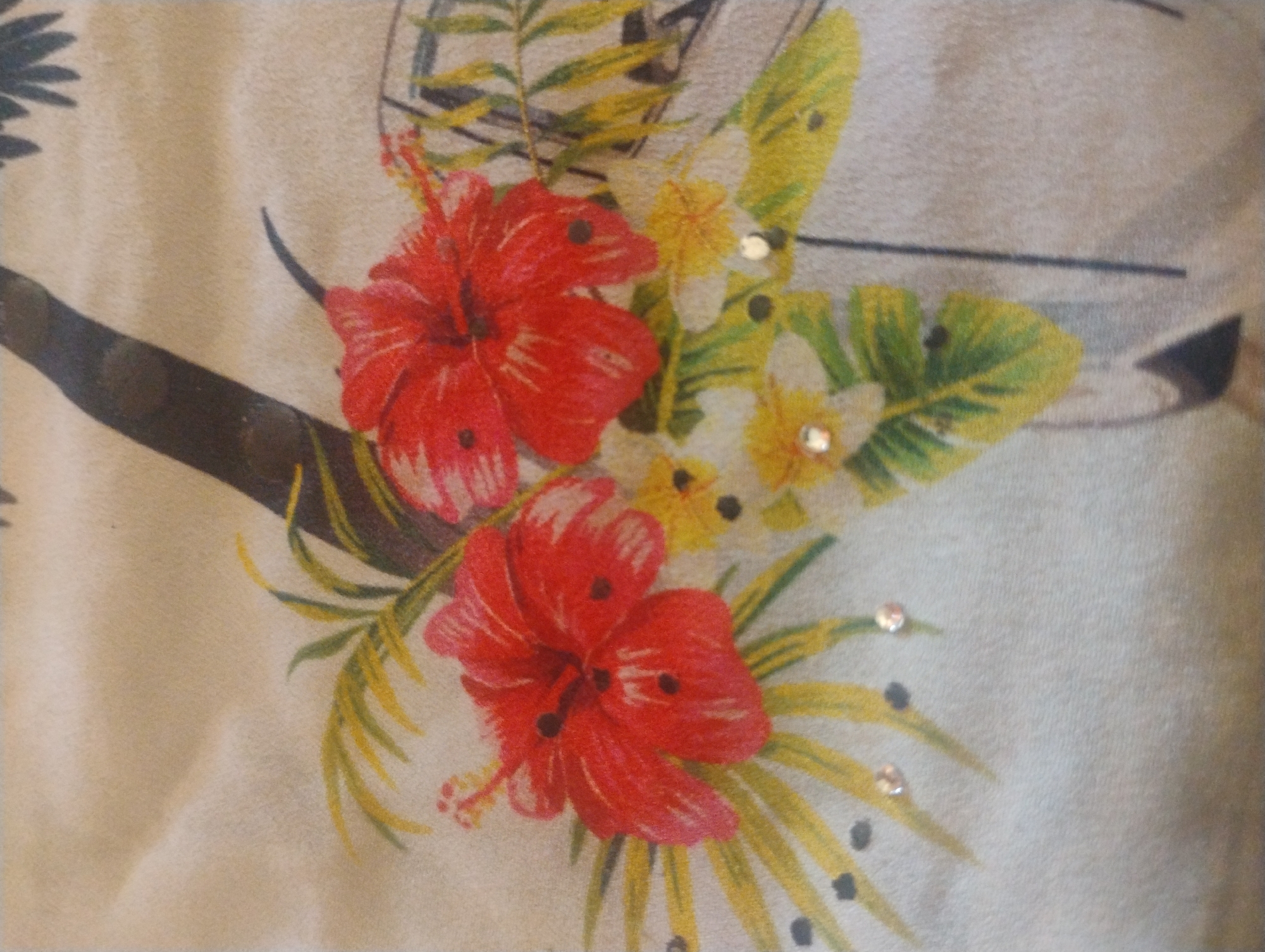 The image shows a close-up of a fabric with a floral print. The print features two prominent red hibiscus flowers with dark centers. Interspersed are smaller yellow and white flowers, as well as green and yellow palm-like leaves. Some of the leaves have small black dots, and there are scattered small, sparkling embellishments that appear to be sequins or rhinestones on the fabric. The background is a light, neutral color, likely beige or cream, with subtle folds and texture from the fabric. The image does not provide any information or context related to the city of Rumyantsevo, Bulgaria.