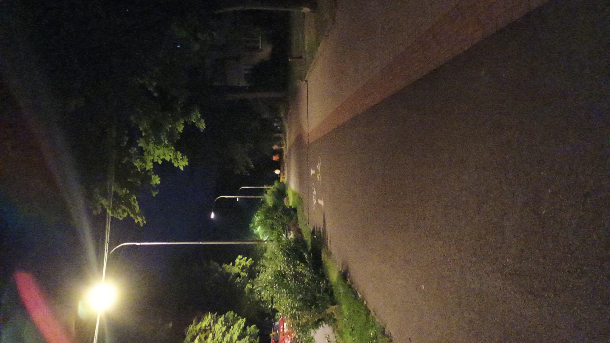 The image depicts a street scene at night in Sokolov, Czechia. A paved pathway, possibly a bike lane, runs through the frame, illuminated by streetlights. To the left, lush green bushes and grass are visible, with a red car parked in the distance. Further down the pathway, several streetlights line the route, and a few individuals can be seen walking in the background, their figures silhouetted against the artificial light. Trees with green foliage flank the path, their branches partially obscuring the dark sky. The overall ambiance is one of a quiet, urban night, with the streetlights casting a warm glow on the surroundings.