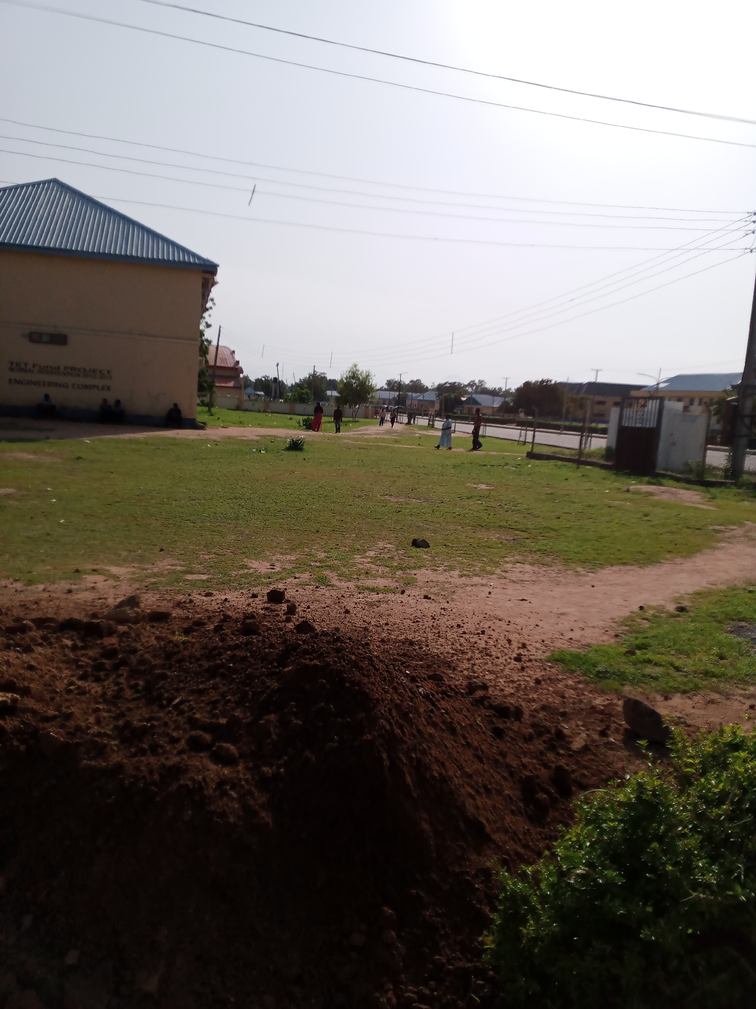 The image depicts an outdoor scene in Jalingo, Nigeria, likely on the grounds of an educational institution. In the foreground, a mound of dark soil is visible, suggesting recent construction or landscaping. Beyond the soil is a well-maintained grassy area, dotted with a few scattered stones.
To the left of the image, a building with a light-colored facade and a corrugated metal roof is partially visible. Text on the building's wall reads "TET Fund Project" and "Normal Intervention 2012-2013 Engineering Complex." Several people are seated or standing near this building.
Further into the scene, a road runs horizontally, with several other buildings visible on the opposite side. These buildings appear to be part of the same complex or a neighboring one. A metal gate is also present along the road.
A few individuals are walking on the grassy area and along the road, engaged in various activities. One person in a red garment is walking away from the camera on the grass. Another pair of individuals, one in a light blue garment and the other in dark clothing, are interacting near the road.
Overhead, power lines are strung across the sky, a common sight in urban and semi-urban environments in Nigeria. The sky is bright and clear, indicating daylight. The overall atmosphere suggests a typical day at a public institution in Jalingo.