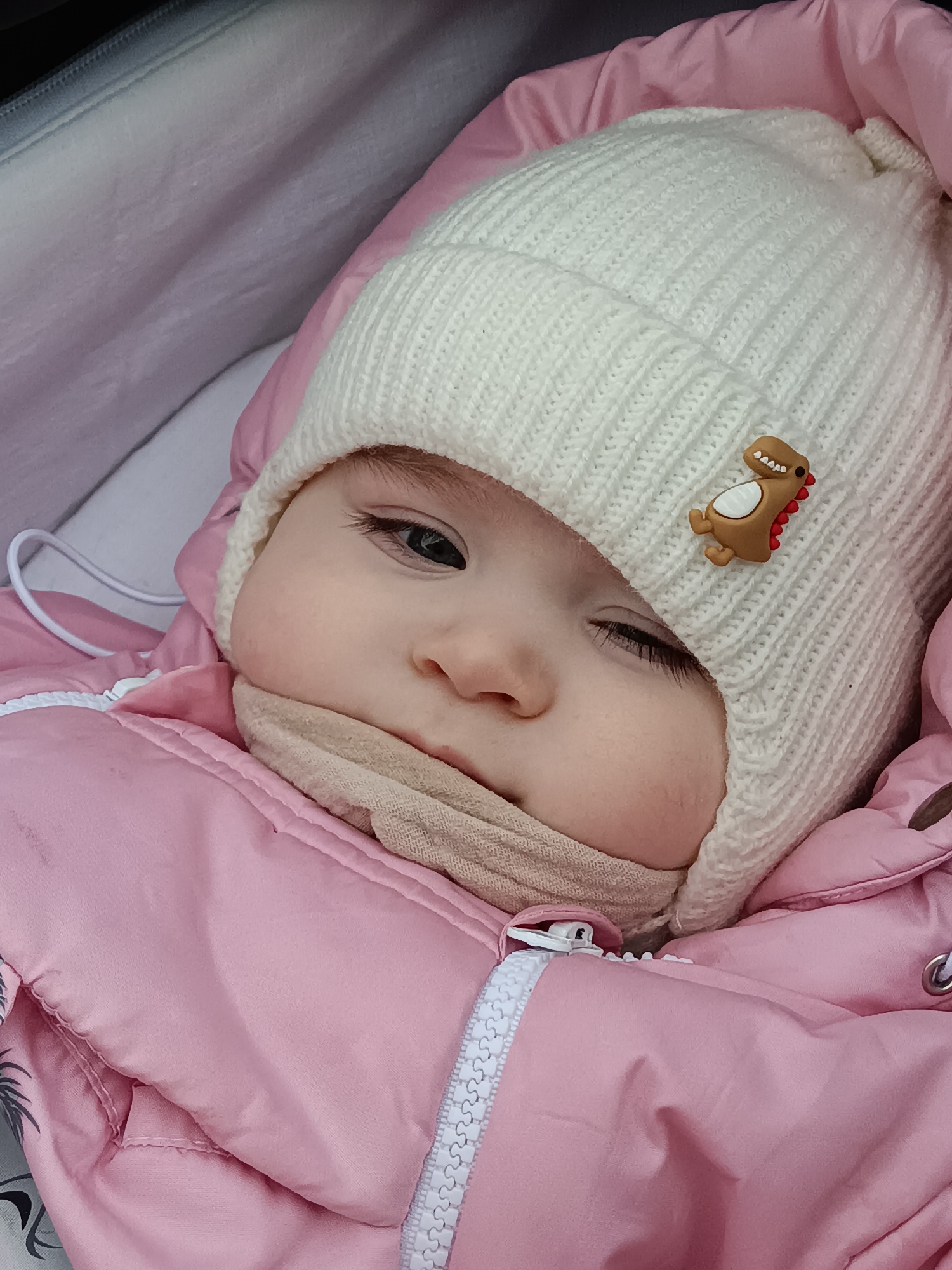 A close-up image captured in Noviy, Russia, displays an infant lying within what appears to be a stroller or pram. The infant is wearing a cream-colored, ribbed knitted beanie hat adorned with a small, brown cartoon dinosaur pin featuring a white belly and red spines. A portion of the infant's face is visible, with one dark eye open and partially shadowed by the hat's brim; the other eye is obscured. The infant's nose and part of the cheek are visible, and a beige knitted neck warmer or scarf is positioned below the chin, obscuring the mouth. The infant is clad in a pink padded winter coat or snowsuit with a white zipper closure visible on the left side of the frame. The immediate environment consists of the stroller's light-colored lining and additional pink fabric from either the stroller or an internal blanket. No other individuals or direct interactions are visible.