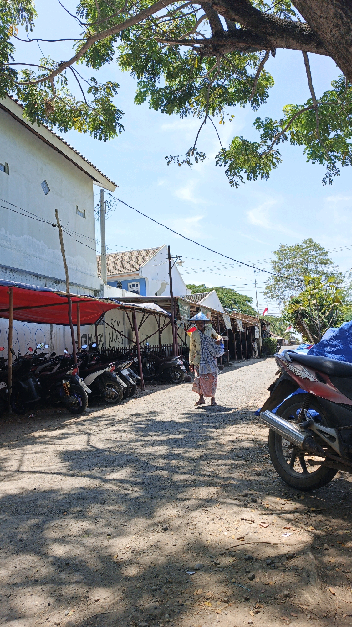 The image depicts a street scene in Krimun, Indonesia, characterized by a rough, gravelly road. On the left side of the street, a row of parked motorcycles is visible, partially covered by a red tarp. A white building with a light blue roof section stands behind the motorcycles, with a utility pole and overhead wires in front of it. Further down the street, a market-like area with stalls and more motorcycles is present. A person, wearing a conical hat and traditional clothing, walks down the street carrying a bundle slung over their shoulder. Trees with green foliage overhang parts of the scene, casting dappled shadows on the ground. The sky is blue with scattered white clouds, indicating daytime. The overall impression is that of a local street with everyday activity.