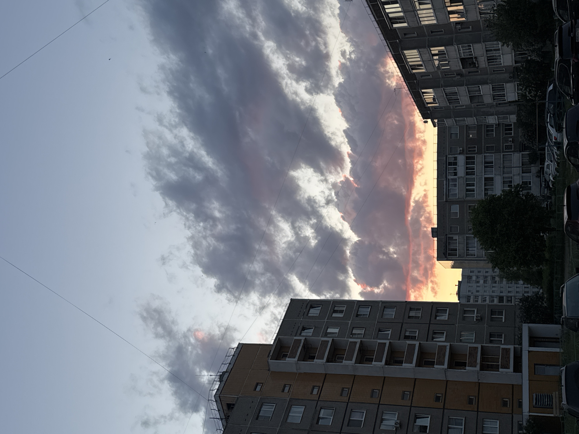 The image depicts an urban scene in what appears to be a residential area. The sky is filled with dramatic, layered clouds, illuminated by a sunset glow of pink and orange, contrasting with darker gray clouds. High-rise buildings dominate the foreground. These buildings feature repetitive window patterns, characteristic of Soviet-era architecture. Several cars are parked along the side of the building. The absence of human subjects suggests a quiet, still moment. The location is presumed to be in Chaikovsky, Russia, based on the prompt.