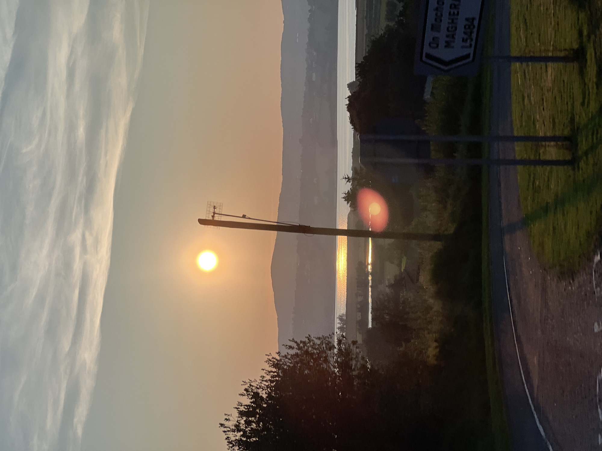 This is a photograph taken at sunset in Letterkenny, Ireland. The image captures a scenic view of a body of water, likely Lough Swilly, with hills in the background. The sun, a bright orb, is setting in the distance, casting a warm glow across the sky and reflecting on the water. In the foreground, a road curves to the left, with a grass verge and some foliage. A signpost is visible on the right side of the road, with text that appears to read "On Machera MAGHERA L5484." There is a telephone pole with an antenna next to the signpost. The overall atmosphere of the image is tranquil and serene, conveying the beauty of the natural landscape at dusk.