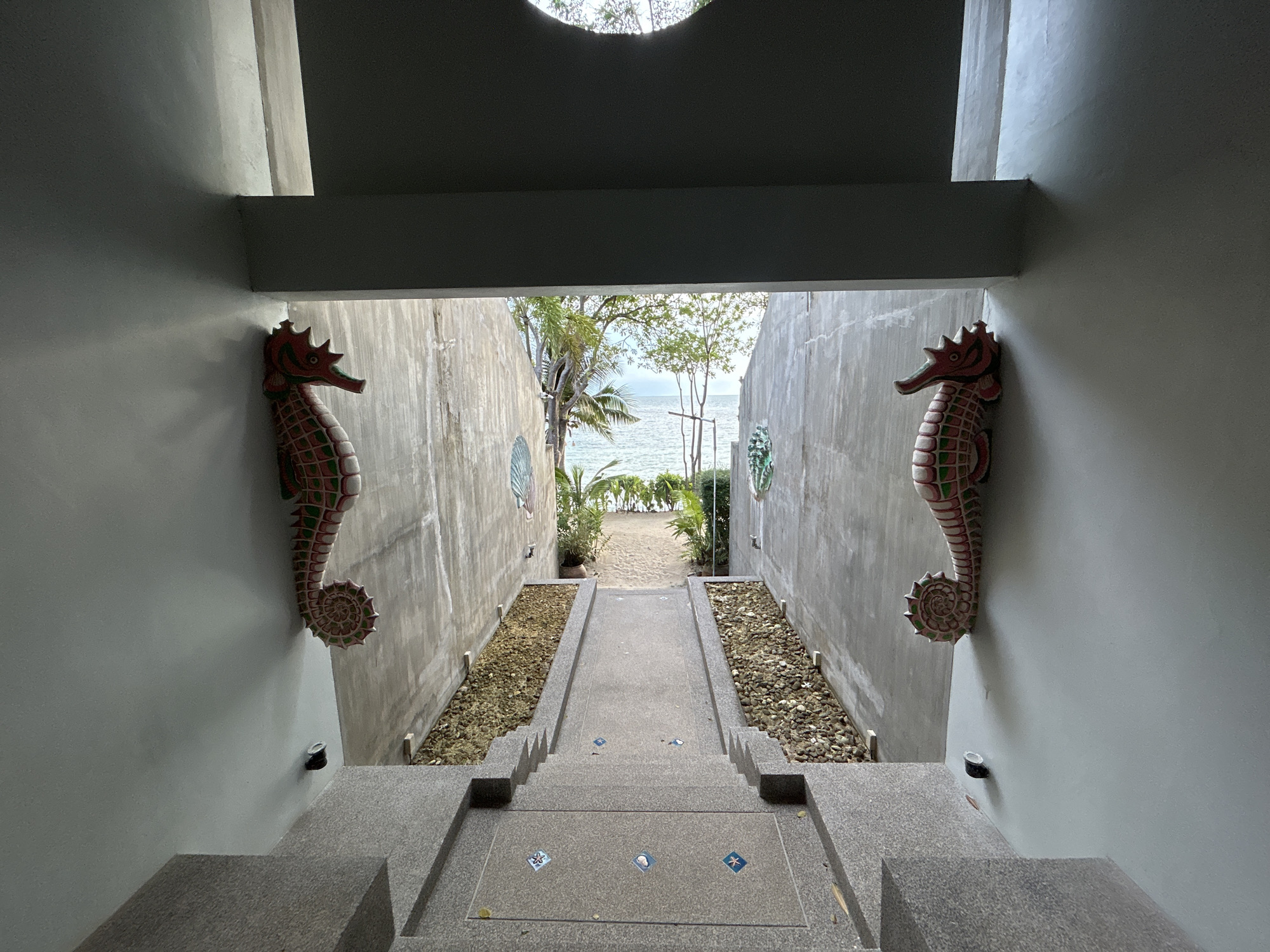 The image depicts a stairway leading to a beach in ตำบลเกาะพะงัน, Thailand. The stairway is constructed of gray stone with blue and white tile inlays. On either side of the stairway are concrete walls adorned with red and green seahorse statues. Small black light fixtures are also visible on the walls. Beyond the walls is vegetation, sand, and ocean water. Trees line the beach. The sky is overcast.