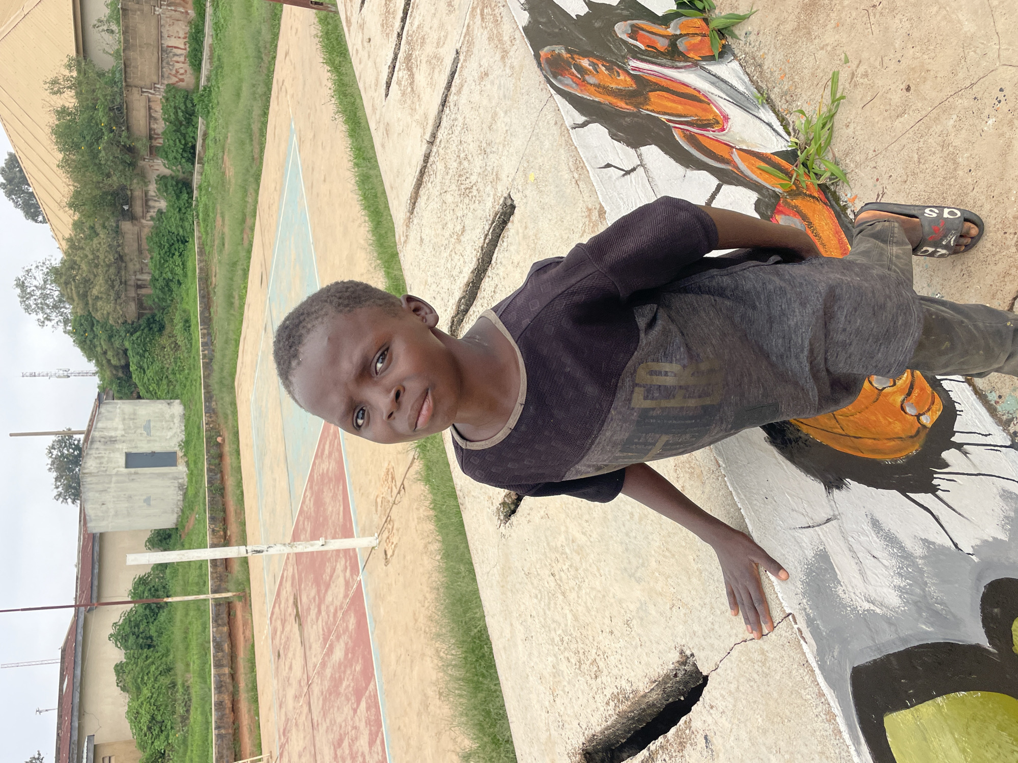 In Gura Top, Nigeria, a young Black boy stands in the foreground, looking towards the camera with a neutral expression. He is wearing a dark, short-sleeved t-shirt with a lighter-colored stripe across the chest and dark pants. The boy's left hand rests on a concrete barrier, which is partially cracked and features a mural of a basketball player in orange and white. His right foot is visible, wearing a black slide sandal with red lettering.
Behind the boy, a red and blue court, likely for volleyball or a similar sport, is visible with a white net pole. In the background, there are low-rise buildings with tan and light gray facades, surrounded by green vegetation. The sky is overcast. The scene appears to be an outdoor recreational area within a settlement.