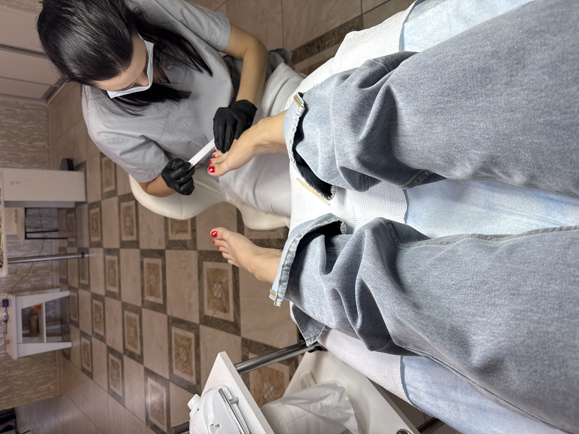 In a well-lit indoor setting, possibly a pedicure salon in Minusinsk, Russia, a person is having their toenails filed. The individual's legs are clad in light-wash denim jeans. The person performing the service, wearing a grey uniform, black gloves, and a face mask, is carefully filing the toenails of the client's right foot. The client's right foot, with bright red nail polish, is positioned for the procedure. To the left of the frame, a white pedicure device rests on a white table. The floor is tiled with a diamond pattern in beige and brown. The background shows two white tables, one with shelves, suggesting a retail or service area.