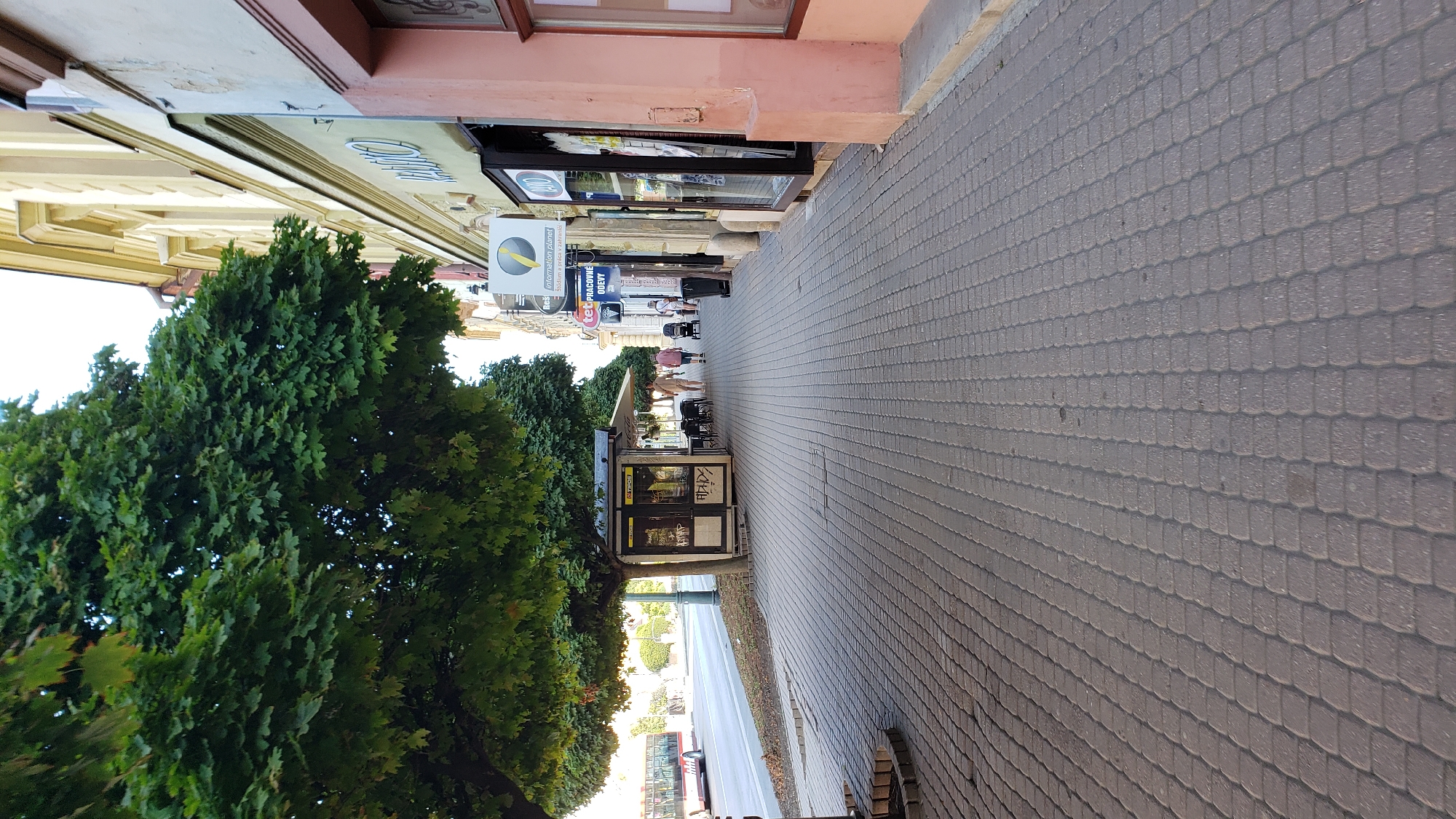 The image depicts a street scene in Prešov, Slovakia, likely on or near Hlavná ulica, a prominent pedestrian street. On the left, a large, leafy green tree shades a sidewalk paved with interlocking gray bricks. Adjacent to the tree is a small kiosk with advertisements on its windows. Further down the sidewalk, a few people are walking, including a couple and a person pushing a stroller. Several shops with large display windows line the right side of the street, featuring various signs and merchandise. A yellow and blue sign with the text "information planet" is visible on one of the buildings. A red bus is visible in the background on the street. The overall atmosphere suggests a sunny day in a European city.