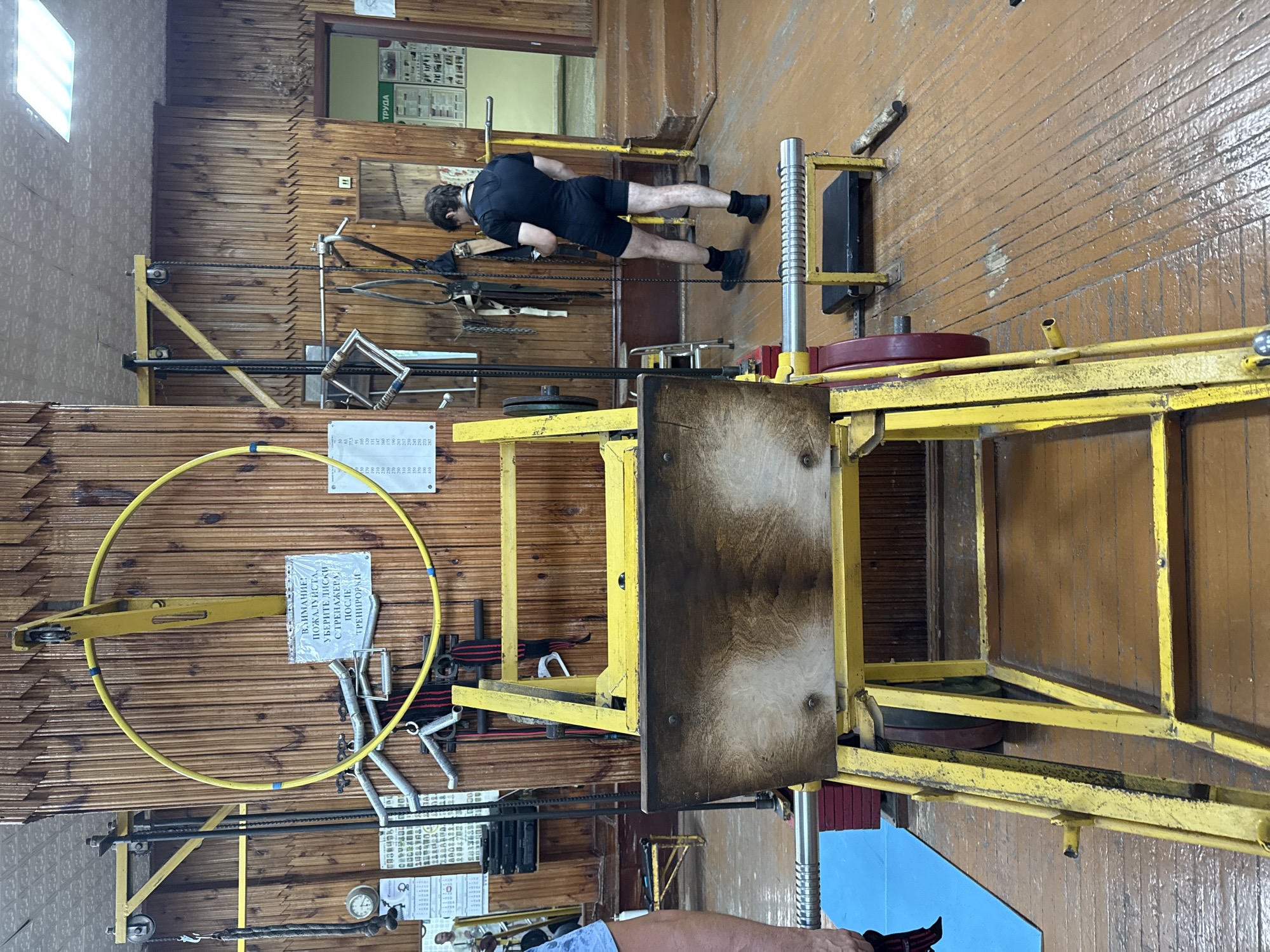 The image depicts the interior of a gym, likely located in Kazan, Russia, characterized by its rustic wooden walls and floor. In the foreground, a yellow weightlifting machine, possibly a leg press, is prominently featured. The machine has a worn wooden platform and metal components. A person, dressed in black athletic wear, stands to the right, facing away from the camera and appearing to engage with a mobile device or watch. The background reveals additional gym equipment, including weights, cables, and a large yellow hoop suspended from the ceiling. A sign with Cyrillic text is affixed to the wall, likely providing instructions or warnings. The overall environment suggests a functional, perhaps older, fitness facility.