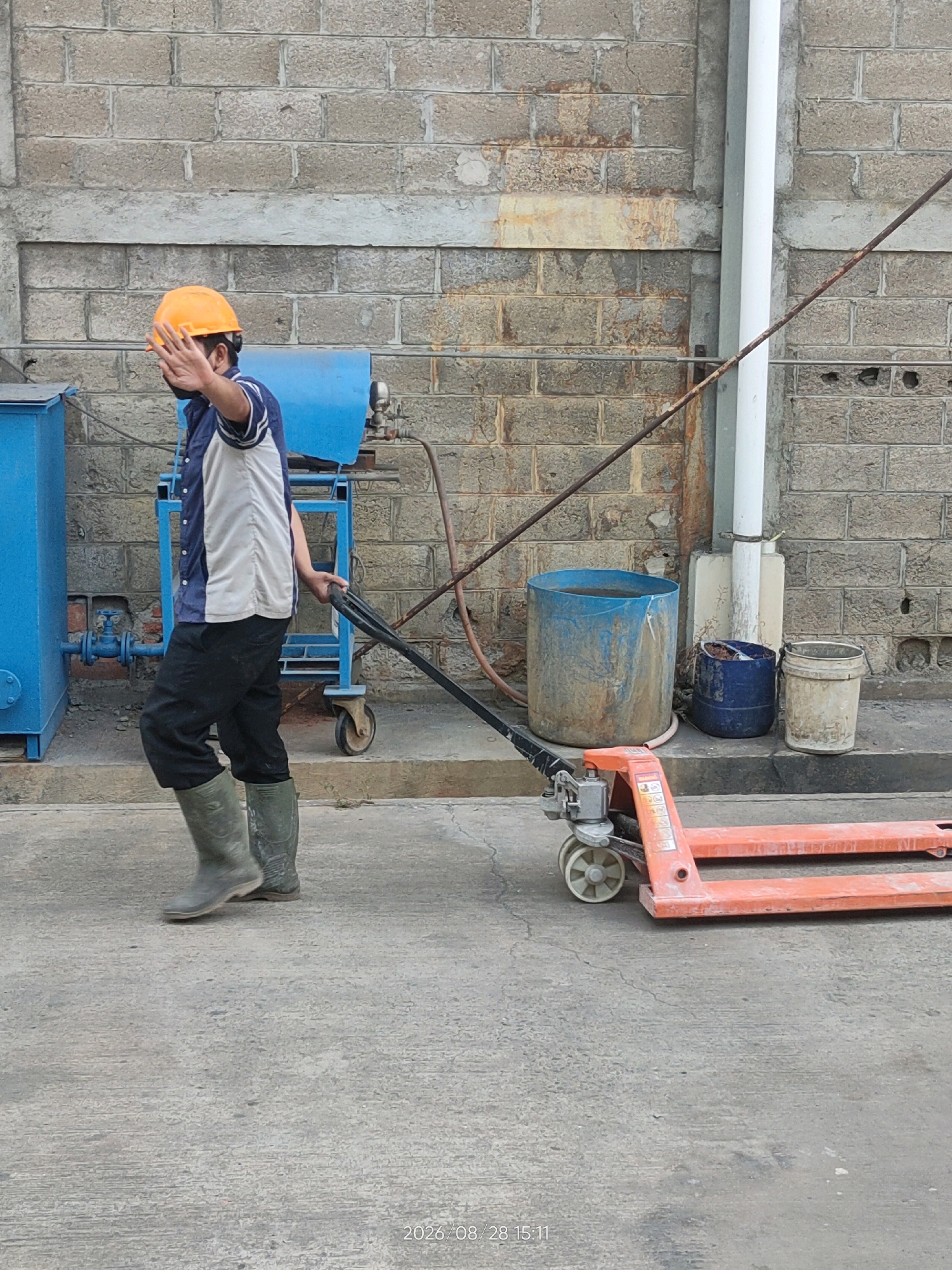 In Telagaasih, Indonesia, a man wearing a hard hat, a short-sleeved shirt with a blue and white pattern, and black pants is seen pushing an orange pallet jack. He is also wearing green rubber boots. The man has his right hand raised, palm facing forward, in a gesture that could indicate stopping or waving. He is walking on a paved surface in front of a brick wall. To his left, there is a blue metal structure with pipes and valves. Behind him, to the right, are several large plastic containers, one blue and two lighter colored. A white pipe runs vertically along the brick wall. The scene appears to be an industrial or storage area.
