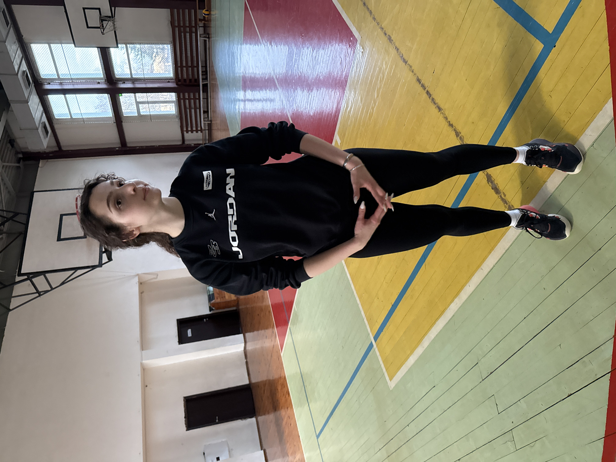 A young adult female stands centrally within an indoor sports hall in Sofia, Bulgaria. She has dark, curly hair, and is looking slightly right and upward with a neutral expression. She wears a black long-sleeved sweatshirt with "JORDAN" printed in white on the chest, black leggings, white ankle socks, and black athletic shoes with pink/orange accents. Her hands are clasped in front of her waist, fingers intertwined. On her left wrist are multiple silver-colored bracelets, and her fingernails are painted white.
The sports hall features a multi-colored wooden floor with sections painted light green, yellow, and red, marked with blue and white lines. In the background, two dark brown doors are visible on a white wall to the left, with a white electrical box mounted nearby. To the right, large windows with a grid pattern allow natural light, showing bare tree branches outside. Below the windows, wooden benches line the wall. A basketball hoop with a white backboard and rim is visible on the right, and another backboard structure is partially visible on the far left. A yellow volleyball is partially visible on the floor near the benches. Overhead lighting fixtures are present near the ceiling. The overall environment is brightly lit, indicative of a gymnasium setting.