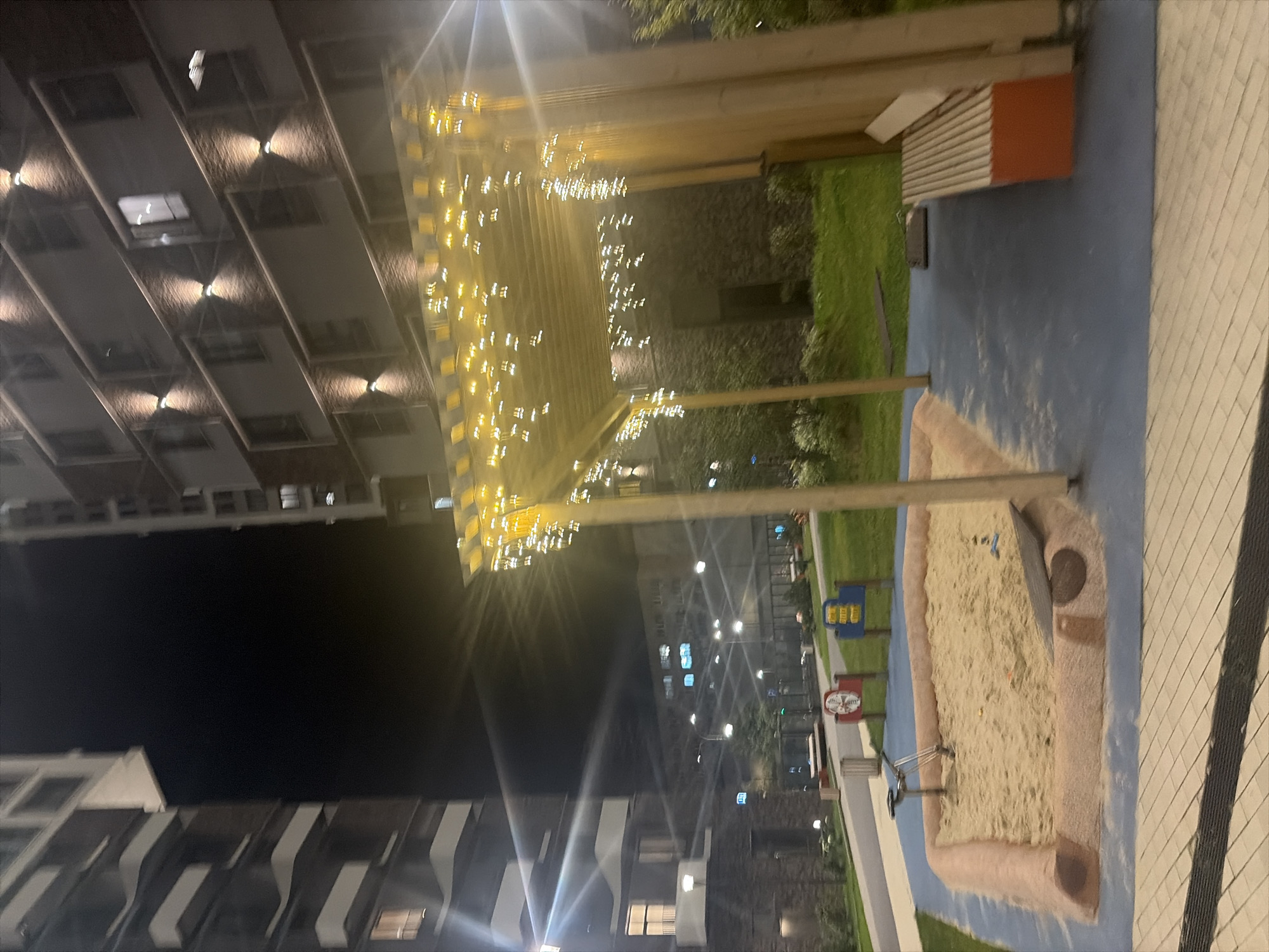 This image depicts a modern playground area at night, likely in a residential complex in Moscow, Russia. The scene is illuminated by ambient light from the surrounding buildings and decorative string lights. In the foreground, a sandbox with a sandy surface is visible, bordered by a light brown material. A small metal swing set is placed within the sandbox. To the right of the sandbox, there is a paved area with a wooden bench that has an orange base. Above the sandbox and bench, there is a wooden canopy structure adorned with numerous small, yellow string lights, creating a festive atmosphere. In the background, tall residential buildings with multiple floors and lit windows rise into the dark sky. Streetlights and other outdoor lighting are also present, casting a soft glow on the green grass and paved pathways. The overall impression is a well-maintained, contemporary urban outdoor space designed for recreation.