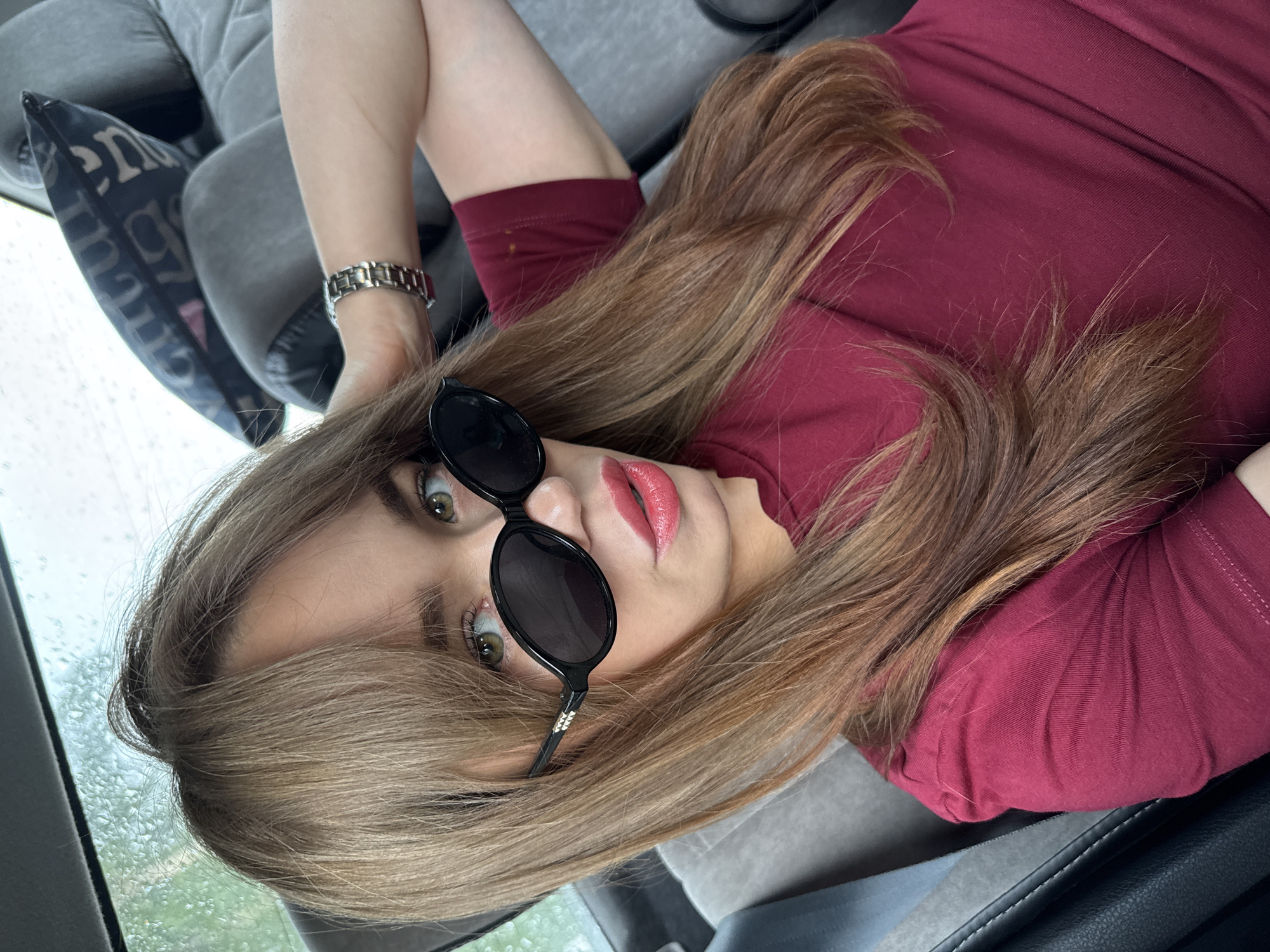 A woman with long, reddish-brown hair and green eyes, wearing sunglasses and a maroon t-shirt, is in the back seat of a car. The car's interior is visible, with grey seats and a headrest pillow with a black pattern. Rain can be seen on the car window. The woman is looking at the camera with her right hand resting on the back of her head, and her left arm is bent and resting on her chest. She is wearing a silver watch on her left wrist. The image appears to be taken in Chaikovsky, Russia.