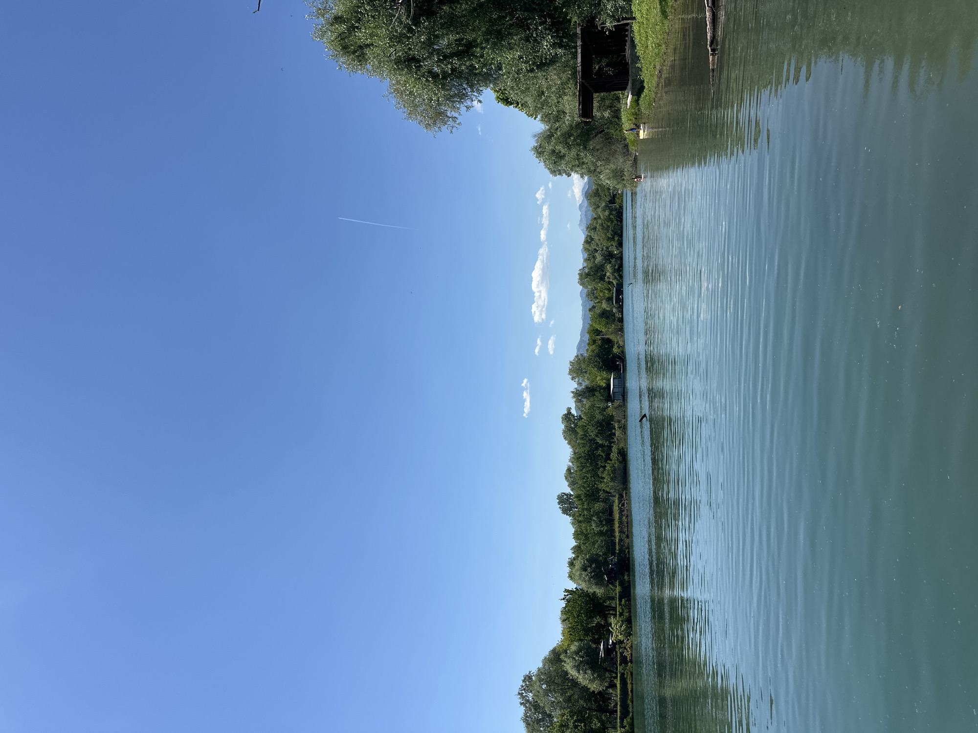 The image shows a body of water, likely a river or lake, flanked by trees and vegetation. The sky is clear blue, with a contrail from an aircraft visible. Along the far bank, there are hints of buildings or structures amongst the trees. The image appears to be taken in Vranjina, Montenegro, based on the geographic features of the landscape, but no identifying markers confirm the exact location. There are no people visible. The camera angle is oriented vertically.