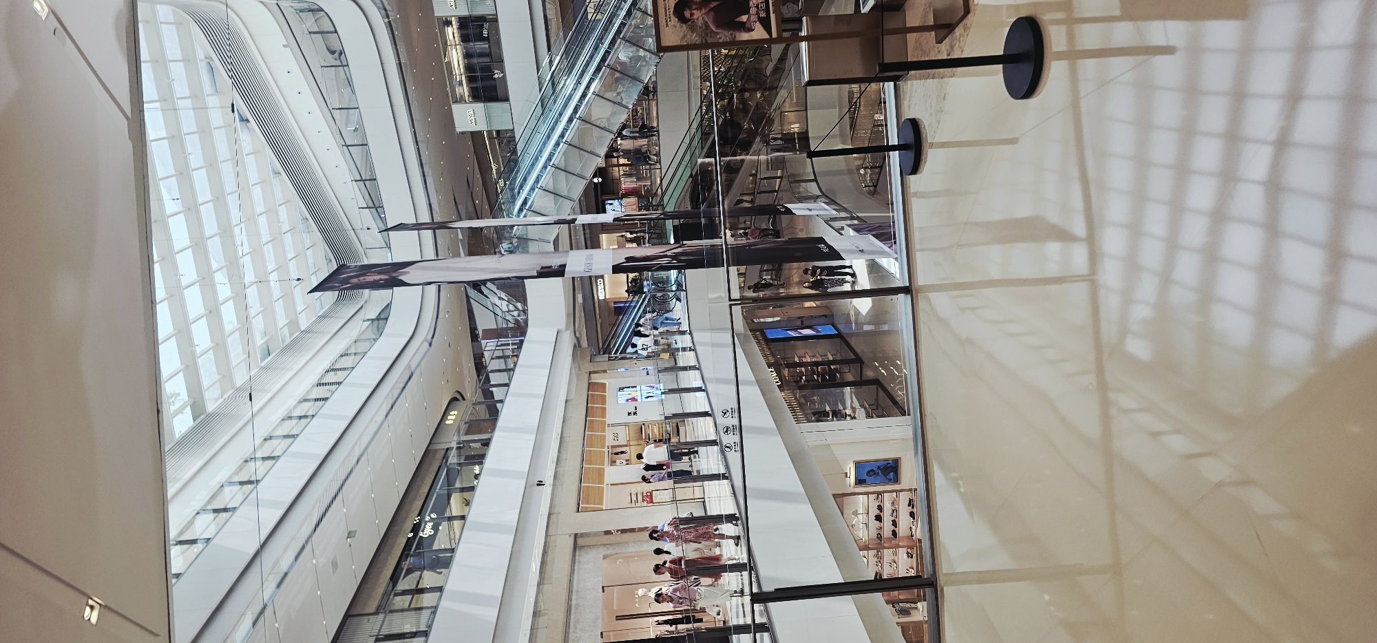 The image depicts the interior of a multi-level shopping mall with a bright, modern architectural design. The scene is viewed from an elevated perspective, looking down into the mall's atrium and onto several floors. Sunlight streams in from a large, angled glass ceiling, illuminating the polished beige tiled floor and reflecting the architectural elements.
On the upper levels, shops with glass storefronts are visible, displaying merchandise and mannequins. Several people are walking along the walkways and escalators. Two large vertical banners featuring a woman's portrait hang in the atrium, adding a touch of commercial branding.
The lower levels show more shop fronts, some with illuminated displays and shelves stocked with items, possibly shoes given the racks seen in one shop. People are scattered throughout the different levels, engaged in shopping or moving between areas. Escalators are a prominent feature, facilitating movement between floors. The overall environment suggests a bustling commercial space. The specific location is identified as a shopping mall in Jiulongpo District, China.