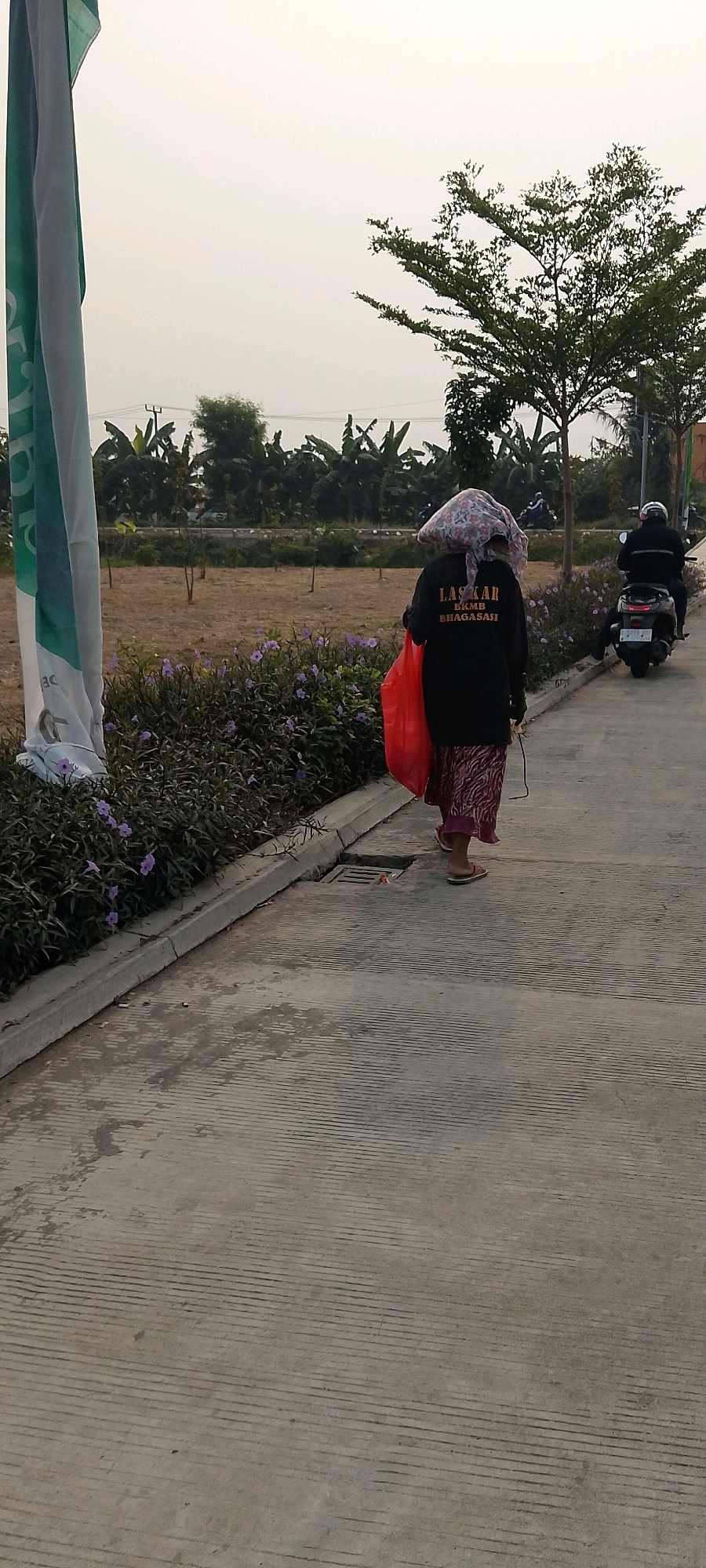 In Sriamur, Indonesia, a person wearing a dark long-sleeved shirt with the text "LASKAR BKMB BHAGASASI" and a patterned skirt walks on a paved pathway. They are carrying a bright red plastic bag in their left hand. The person is also wearing a floral headscarf. To the right, a person on a scooter is riding away from the camera. The environment features a grassy area with scattered trees and banana trees in the background. A green and white flag is visible on the left side of the image. The sky is overcast.