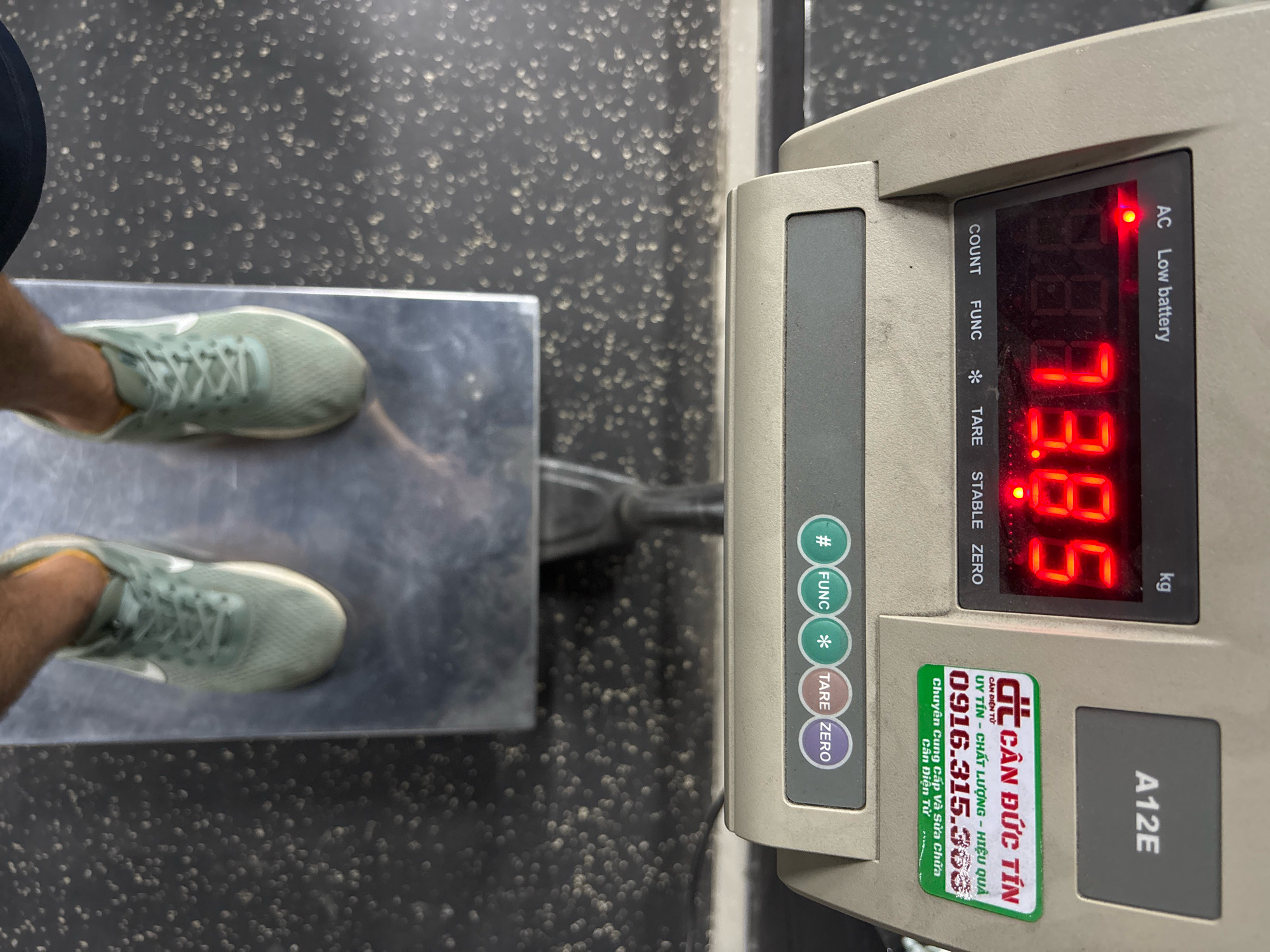The image shows a person standing on a digital scale. The scale displays a reading of 88.8 kg. The person is wearing light green athletic shoes. The scale is located in a room with a dark, speckled floor. A sticker on the scale reads "Cân Đức Tín" and includes a phone number in Vietnamese. The scale is also labeled "A12E". The scale is plugged into an AC power source, indicated by a red light labeled "AC" and text "Low battery". The image appears to have been taken in Nha Trang, Vietnam, based on the Vietnamese text on the sticker.