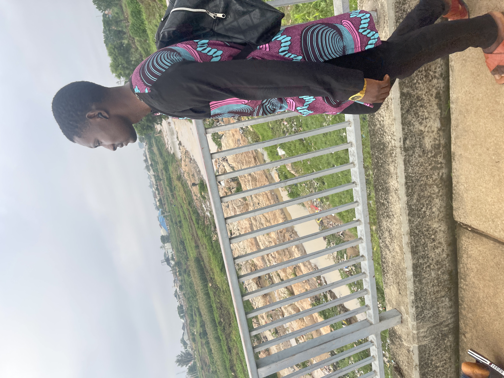 A medium shot captures a young Black man walking along a bridge in Rafin Par, Nigeria. He is looking to his right, away from the camera. He wears a black long-sleeved shirt with a vibrant geometric pattern in pink, turquoise, and black, black pants, and brown sandals. A black backpack is slung over his left shoulder. The bridge railing is made of gray metal bars. Below the bridge, a shallow, muddy riverbed is visible, littered with debris and surrounded by dry, rocky terrain and patches of green vegetation. In the background, a hazy cityscape with scattered buildings, trees, and a large body of water can be seen under an overcast sky. The overall environment appears somewhat neglected, with evident pollution in the riverbed.