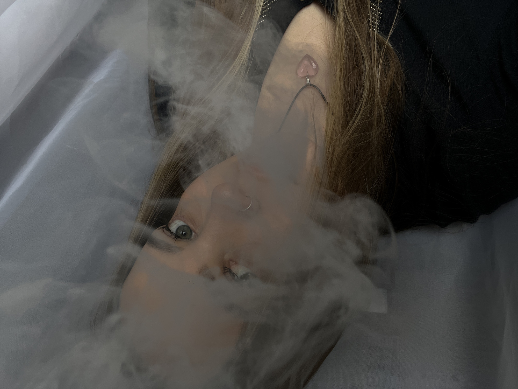 A tilted close-up shot depicts a person with light-colored hair, partially obscured by a white, smoke-like substance. The individual has light skin, visible green eyes, and a silver nose ring. They appear to be wearing a dark top and a necklace with a pink, heart-shaped pendant. The background features a draped, light-colored fabric. The location of Novopokrovskaya, Russia, cannot be determined from the visual data provided.