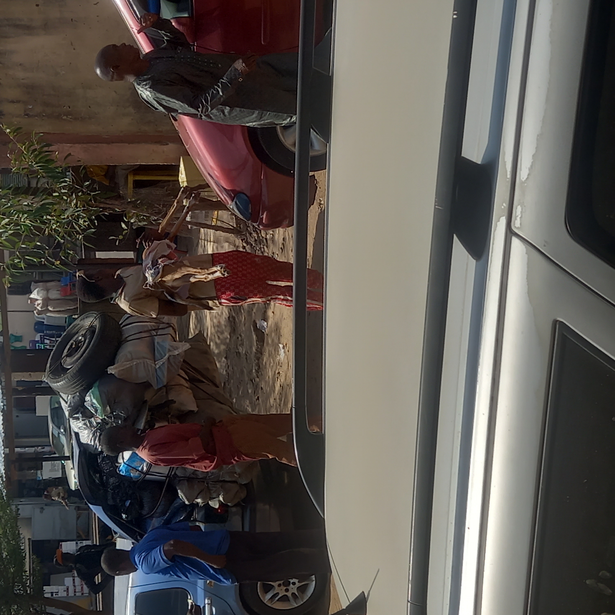 In Sardauna, Nigeria, a scene unfolds near a building with red and beige tones. Several people are present; one man stands partially inside a red car, wearing a dark, patterned garment. Nearby, two other individuals are seen with goods and items, including a tire, loaded onto what appears to be a vehicle or cart. A man in a blue shirt is also visible. The area is filled with vehicles and what seem to be various items and goods, suggesting a busy or market setting. The dominant foreground element is the side of a white car, which seems to be the vantage point from where the image was captured.