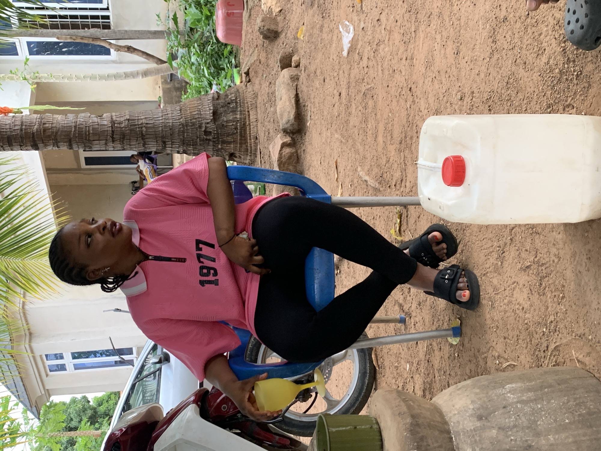 In an outdoor setting in Akwanga, Nigeria, a young woman sits on a blue plastic chair, wearing a pink shirt with "1977" printed on it and black leggings. She holds a yellow container near a red and white motorcycle, apparently pouring something into it. A large white plastic container with a red cap sits to her right on the dirt ground. The ground is dirt. Part of a building with white walls and windows, as well as trees, are visible in the background.