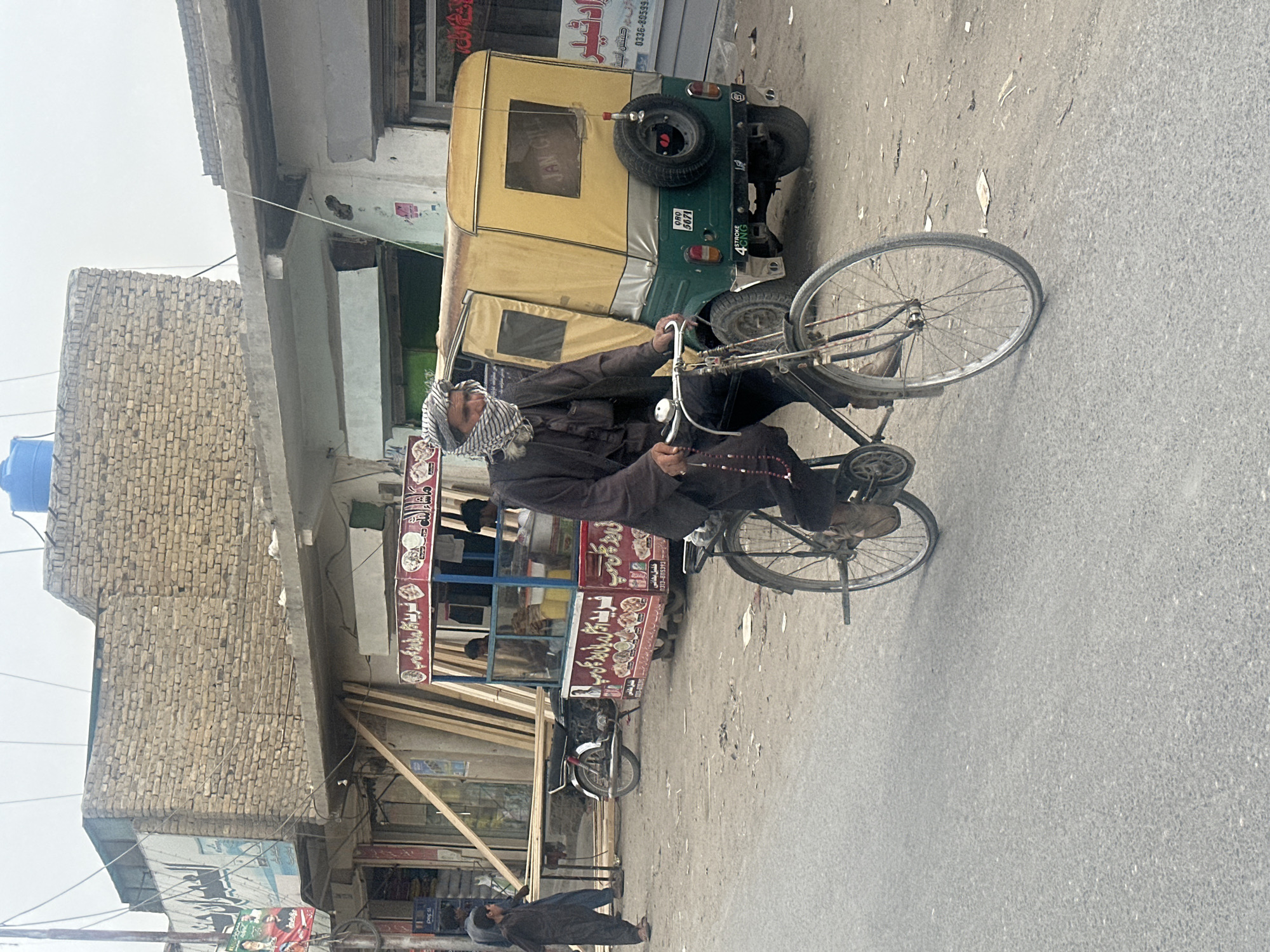 The image captures a street scene in Quetta City Tehsil, Pakistan. A man wearing a headdress and dark clothing is riding a bicycle. Alongside him is a three-wheeled vehicle (tuk-tuk) with a spare tire attached to its rear, parked near a food stall with red and white signage. The building in the background is constructed from light-colored bricks. Other elements include a motorcycle parked next to the food stall, utility poles and power lines, and another person walking on the side of the road. The setting suggests an urban environment, with businesses and vehicles sharing the street space.