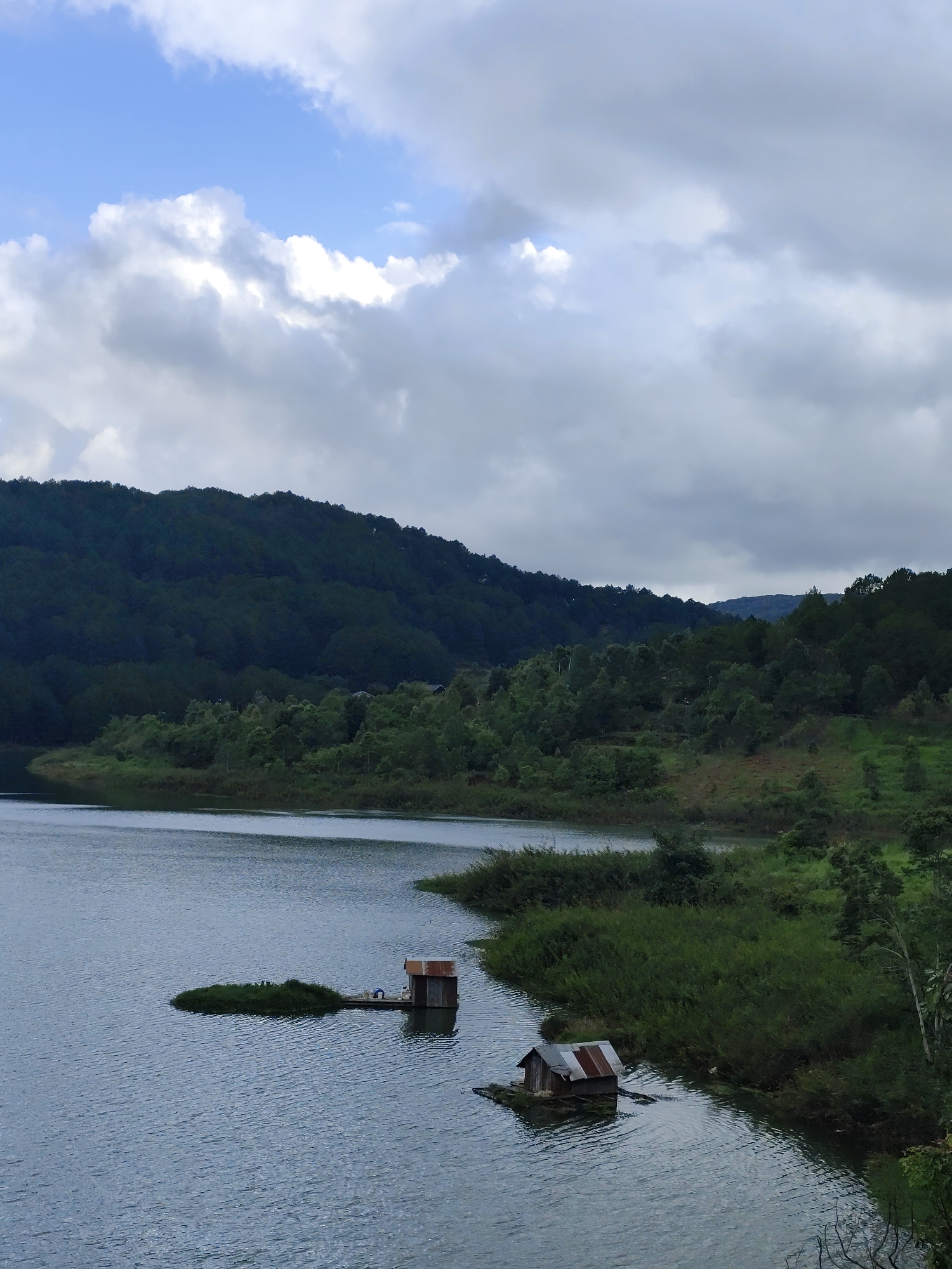 The image depicts a tranquil lake scene in Da Lat, Vietnam. The foreground features the lake's surface with gentle ripples. Two small structures, likely floating shelters or docks, are positioned near the shoreline. One structure includes a small shack with a rusty metal roof. Lush green vegetation lines the edge of the lake. In the background, a forested hill rises, its slopes covered in dense trees. The sky is overcast with patches of blue visible amidst the clouds.