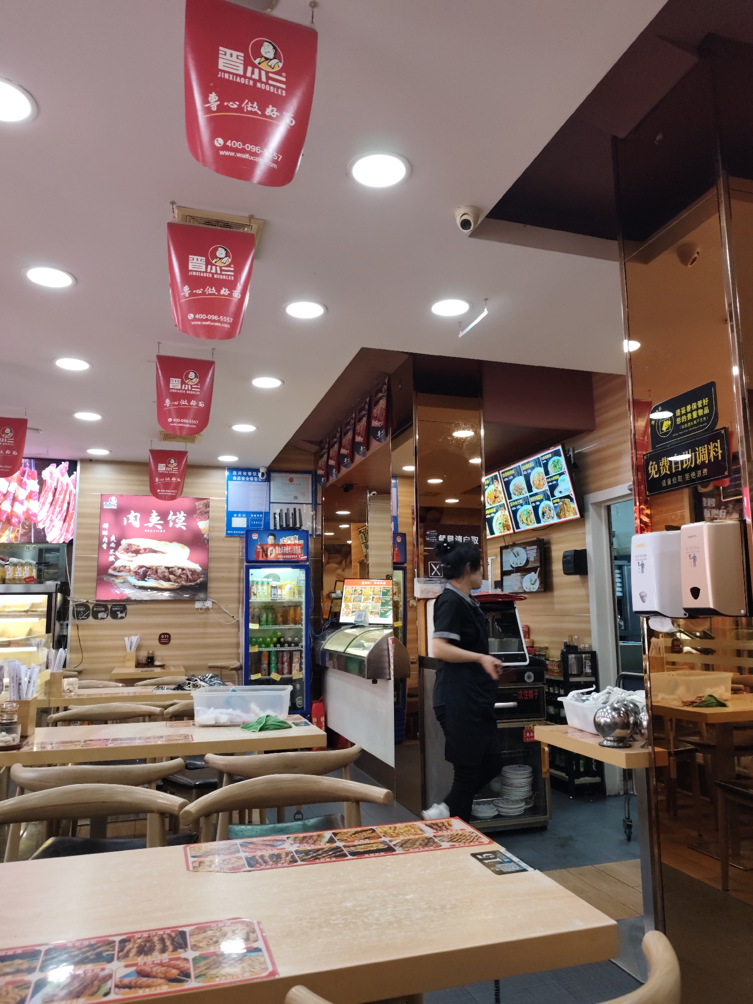 This image depicts the interior of a restaurant, likely a noodle shop, located in Aihui, China. The establishment features a modern design with warm lighting and wooden accents. Several red banners with Chinese characters and branding are suspended from the ceiling. In the foreground, tables and chairs are arranged for dining. A woman in a black uniform, presumably an employee, is visible behind a counter, attending to a point-of-sale system.
The restaurant's menu is displayed on a digital screen, showcasing various dishes. A refrigerator stocked with beverages is also present, along with display cases containing food items. The walls are decorated with posters and signs, including a large advertisement for "JINXIAOER NOODLES" and information about phone numbers and websites. A security camera is mounted on the ceiling. Reflections in a mirrored wall reveal additional details of the restaurant's layout and patrons. The overall atmosphere suggests a bustling, well-lit dining establishment.