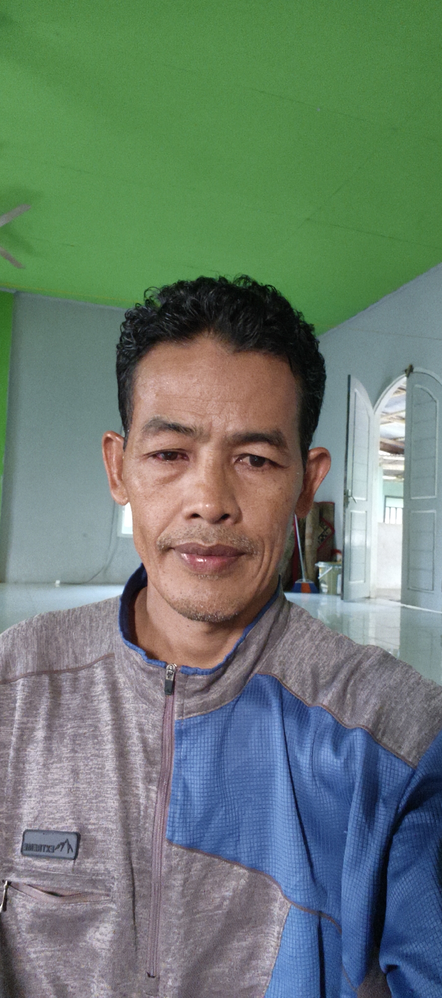 A close-up, waist-up, selfie-style photograph shows a middle-aged Indonesian man in Baru, Indonesia. He has dark, curly hair, a mustache, and a goatee. His skin is tanned and wrinkled, and his eyes appear red and slightly swollen. He is wearing a zipped jacket with gray and blue panels. The background is a room with a green ceiling and light blue walls. Double doors with arched tops are visible in the background, leading to another part of the room. A ceiling fan is partially visible in the upper left corner. The floor appears to be tiled. The image is a selfie, suggesting the subject is taking the photo himself.