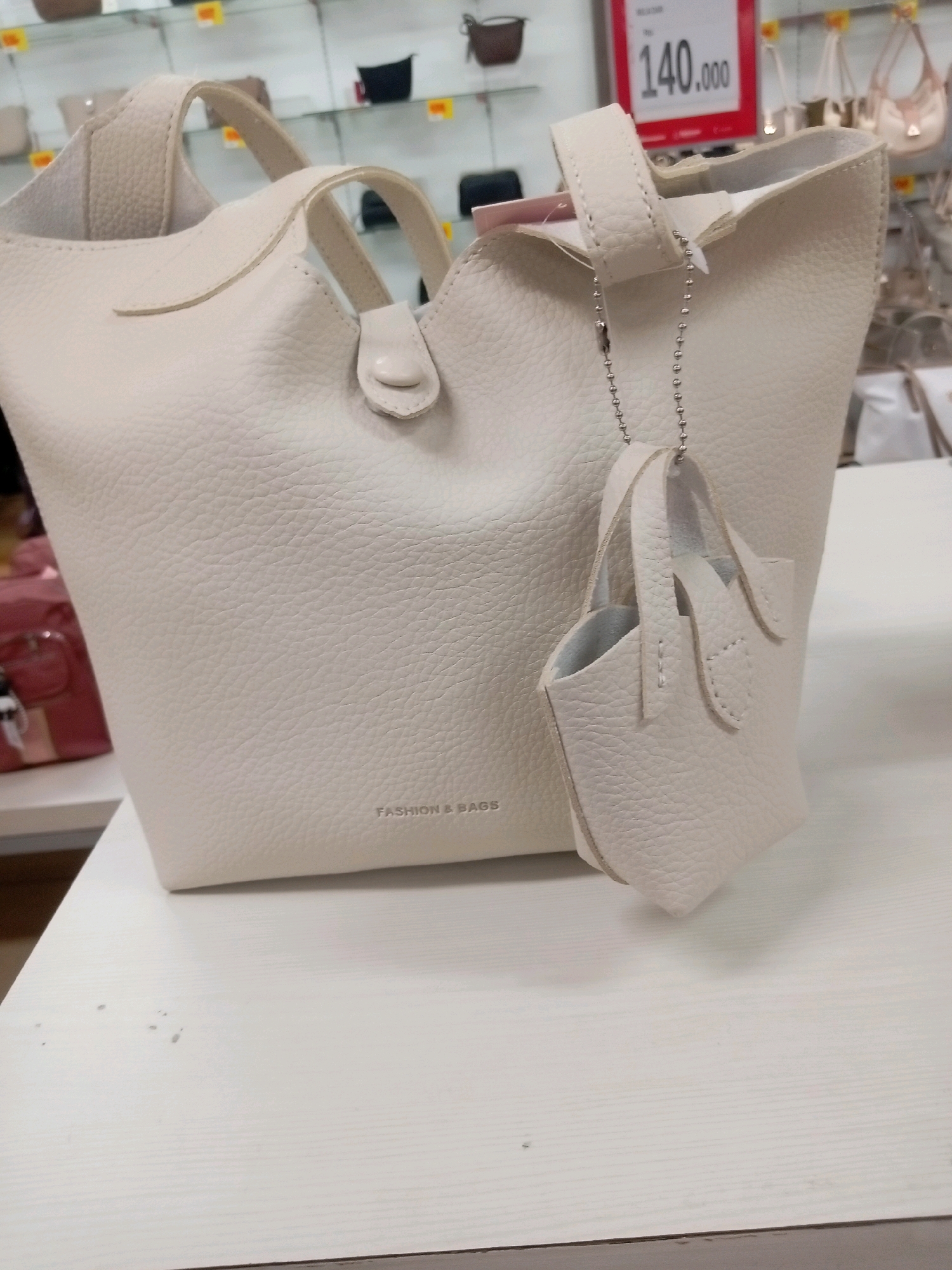 A white, pebbled leather handbag with a small matching pouch attached by a silver beaded chain is displayed on a white, textured surface. The handbag has two top handles and a subtle embossed "FASHION & BAGS" logo on its side. In the background, a red price tag displays "140.000" and other handbags are visible on shelves, suggesting a retail environment. The scene is indoors, likely in a department store or boutique, possibly in Batam, Indonesia, based on the context provided by the query. There are no people or explicit interactions depicted. The lighting is bright, illuminating the handbag and its details.