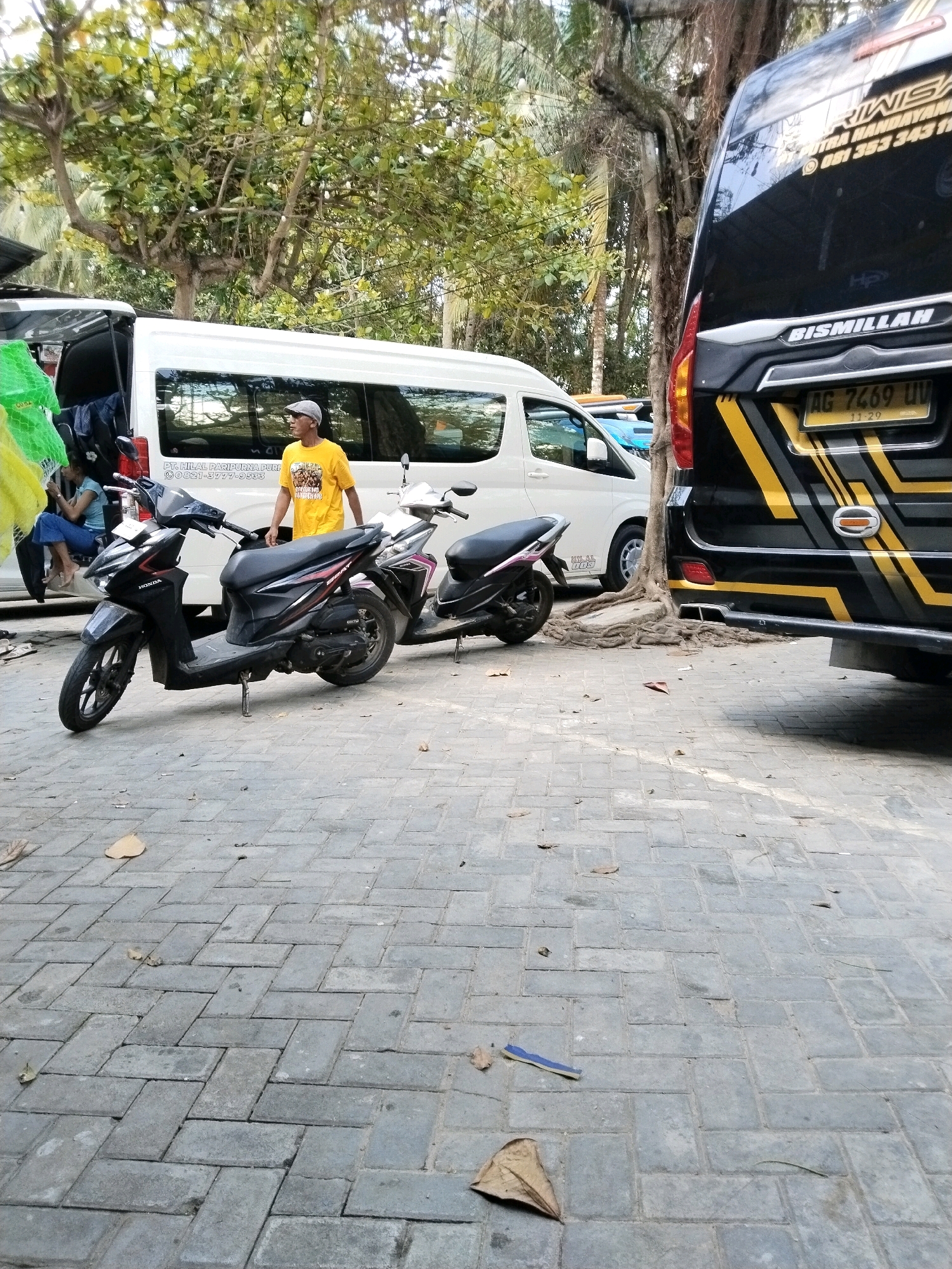 A man wearing a yellow t-shirt and a grey cap stands next to a black Honda Beat scooter, leaning on it. The scooter is parked on a cobblestone pavement. Behind the man and scooter is a white van with tinted windows. To the right of the van, a black bus with yellow stripes is visible, its rear end featuring a license plate with the text "AG 7469 UV". The scene appears to be outdoors, with trees and foliage in the background. The overall setting suggests a transportation hub or a street in a tropical or subtropical region, possibly the city of Prigi, Indonesia, given the information provided.