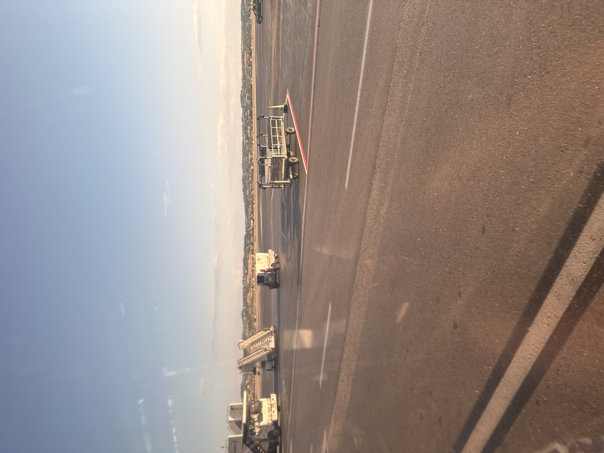 The image depicts an outdoor scene at an airport with an asphalt tarmac under a clear blue sky. In the mid-ground, there are several airport support vehicles and equipment. To the left, a white vehicle with a boarding stairs attached is parked. Further to the right, another smaller vehicle with a boarding stairs is visible, and next to it, a white utility vehicle. In the foreground, a luggage cart with a yellow safety cone is parked. The background shows a distant town or city with buildings and vegetation, likely the city of Thermi, Greece, given the context of the prompt. The overall lighting suggests it is late afternoon or early evening, with the sun casting soft light and long shadows. There are no people visible in the frame.