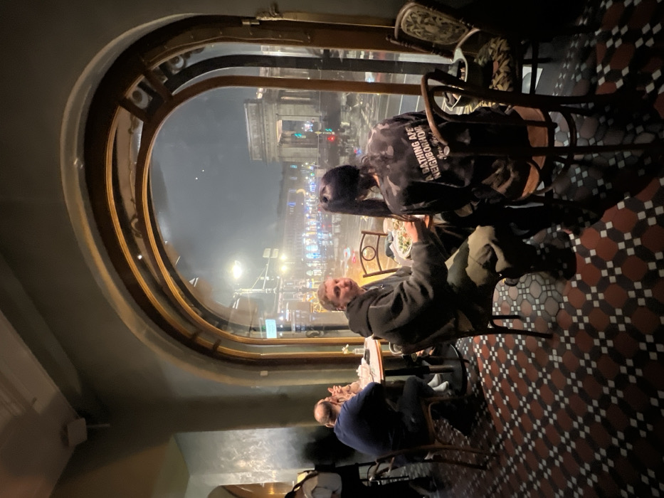 The image depicts the interior of a restaurant or cafe, viewed towards a large arched window overlooking a city street at night in Saint Petersburg, Russia.
**Location:** The scene is set within an establishment on a major thoroughfare in Saint Petersburg, likely Nevsky Prospekt, with a direct view of the Arch of the General Staff Building in the distance, a prominent landmark connecting the General Staff Building's wings and leading to Palace Square.
**People:**
1. **Foreground Left:** A male individual with short, light-brown hair, wearing a dark hooded jacket and camouflage-patterned trousers, is seated at a table. He is turned slightly towards the camera, with his mouth open, and appears to be in the process of eating. A plate of food is in front of him.
2. **Foreground Right:** A second individual, seen from behind, is seated across the table from the male. This person has long dark hair styled in a high ponytail and wears a dark, tie-dye style long-sleeved shirt with the text "A BATHING APE NEIGHBORHOOD New York" visible on the back.
3. **Midground Left:** Two individuals are partially visible at another table. One, a bald man in a dark blue shirt, has his back mostly to the camera and is conversing with another person whose light-colored sleeve and arm are visible across the table.
4. **Far Left:** A figure in a light top and dark pants is partially visible, standing or walking near an interior archway.
**Objects:**
- **Furniture:** Several dark wooden chairs with curved backs, some upholstered, and circular tables with dark bases. One empty upholstered chair is near the foreground window.
- **Dining Items:** Plates of food, appearing to contain salads or similar dishes, are on the foreground table. A small white vase with a single light-colored flower is on the midground table.
- **Interior Structures:** The restaurant features walls painted a muted green-grey, with decorative textured sections. A large arched window with prominent dark wooden or metal framing dominates the view. An air vent is visible on the ceiling in the upper left.
- **Flooring:** The floor is covered in a mosaic pattern of black, white, and red geometric tiles.
**Actions/Interactions:**
- The two individuals in the foreground are seated at a table, presumably dining. The male is looking towards the camera while chewing.
- The two individuals in the midground are seated and appear to be engaged in conversation or dining.
- The far-left figure suggests general movement within the establishment.
**Environment (Interior):** The interior is dimly lit, with artificial light creating reflections on surfaces and the window. The overall ambiance appears to be that of a casual dining establishment.
**Environment (Exterior):** Visible through the large arched window is a busy city street at night. Streetlights and vehicle headlights illuminate the scene. The sky is dark and overcast. Vehicle light trails indicate ongoing traffic. The Arch of the General Staff Building is clearly visible in the distance, illuminated. Other city buildings are visible on either side of the arch. Reflections of interior lights are present on the window glass. Snow or slush is visible on the ground outside, indicating cold weather conditions.