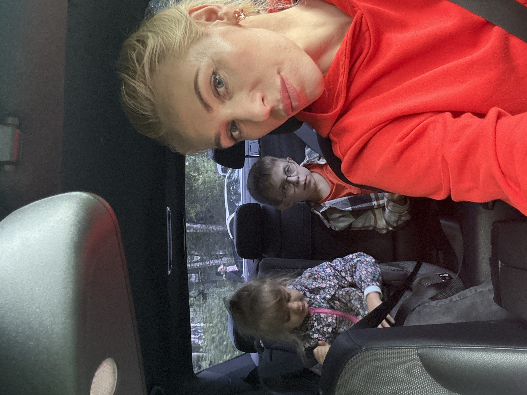 The image captures a close-up, selfie-style shot from inside a car, likely in Saint Petersburg, Russia, based on common aesthetic choices and the general appearance of the surroundings, though no specific landmarks confirm this. A woman with blonde hair, wearing a red hooded sweatshirt and distinctive amber earrings, is prominently featured in the foreground, her gaze directed towards the camera. Behind her, two children are seated in the back of the vehicle. A young girl with brown hair, wearing a floral jacket with a pink zipper, is on the left, appearing to interact with something in her lap. To her right, a boy with glasses and a plaid shirt over an orange t-shirt is visible, looking slightly to the side. The interior of the car is dark, with the car's sun visor visible at the top. Through the car windows, a backdrop of trees and foliage can be seen, suggesting an outdoor, possibly suburban or park-like setting. There are other vehicles visible in the distance, indicating they are on a road or in a parking area. The overall lighting suggests it is daytime.