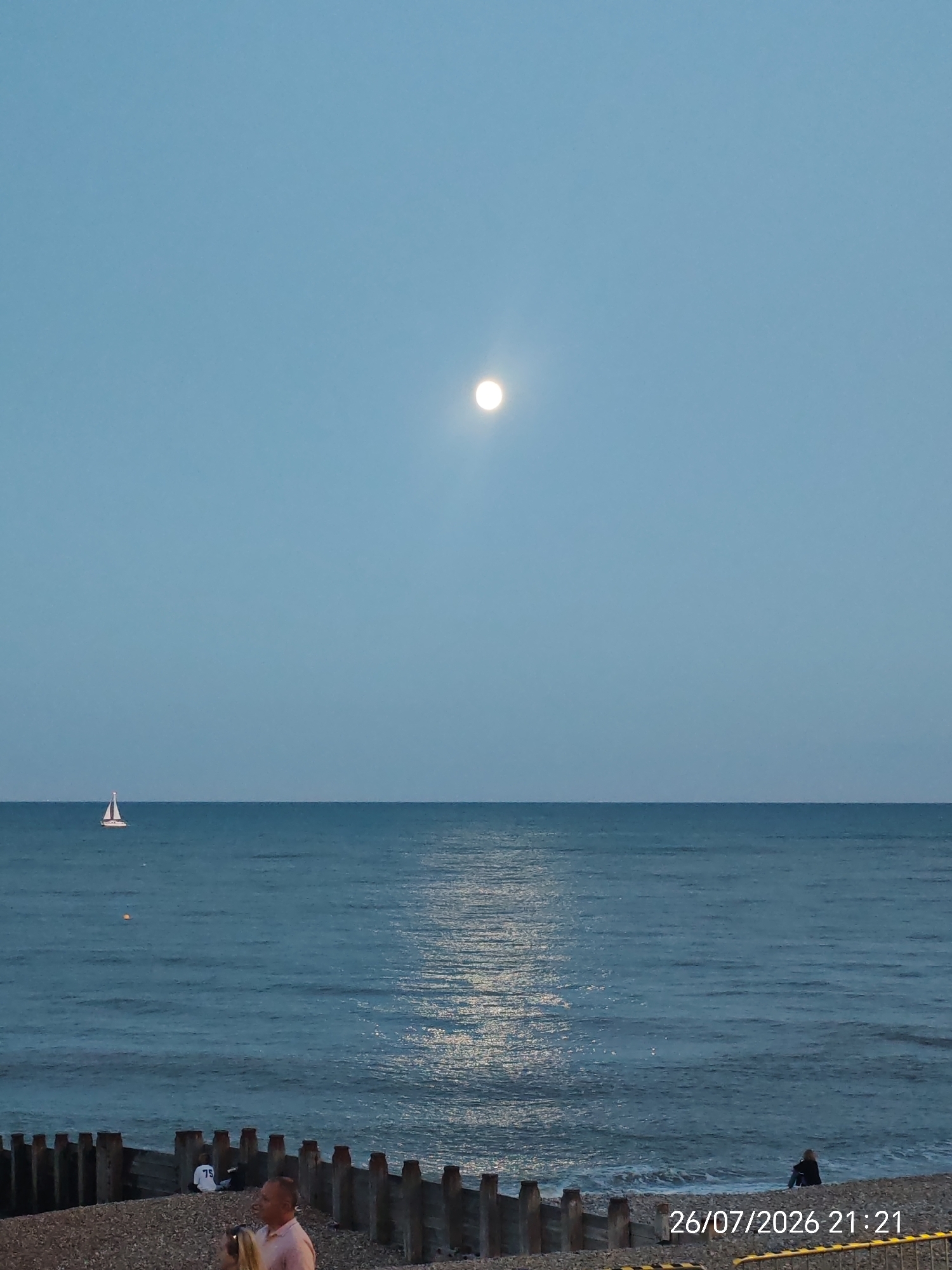 The image depicts a twilight scene at Eastbourne, United Kingdom, with the full moon prominently visible in the clear, pale blue sky. The sea is calm, reflecting the moonlight with a shimmering path. On the horizon, a lone white sailboat with a red hull is visible. The foreground features a pebbled beach with wooden groynes, some of which are partially submerged in the water. Two individuals are present on the beach: one person, dressed in a light pink shirt, is standing in the foreground and appears to be holding a phone, possibly taking a photograph. Another person is seated further down the beach, facing the sea. A date and time stamp "26/07/2026 21:21" is overlaid on the lower right corner of the image.