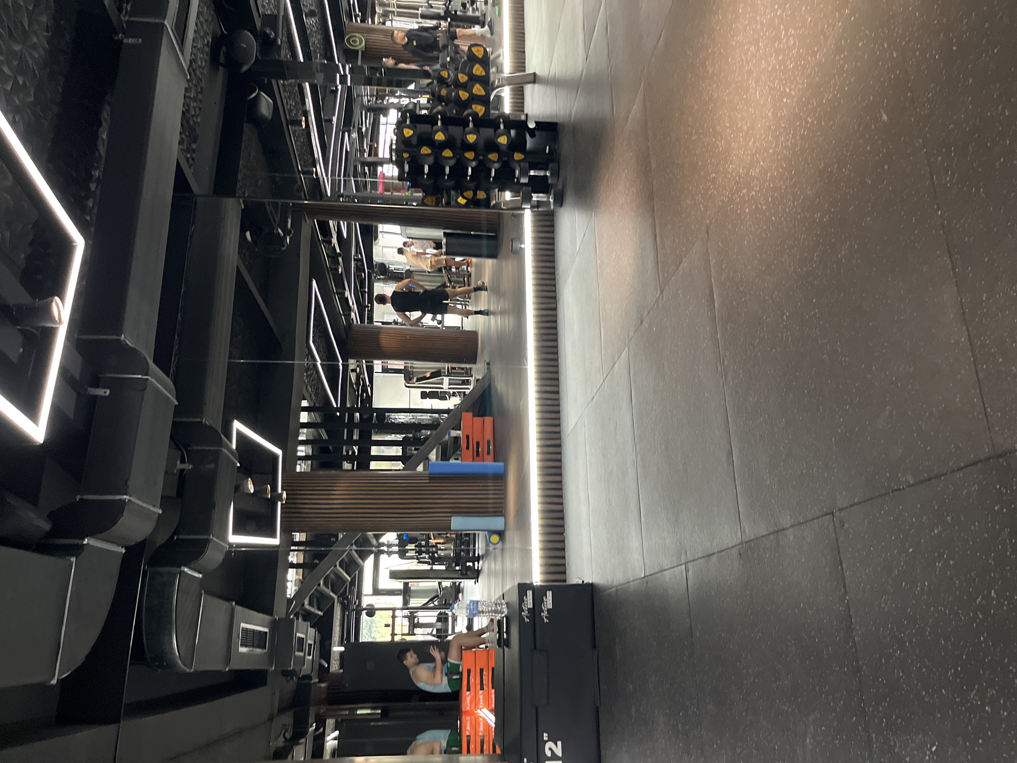 A gym interior features exercise equipment and individuals engaged in physical activity. Dumbbells are arranged on racks, and people are working out. A person is seen performing crunches on a box. The environment includes lighting fixtures and ventilation systems. No definitive information to confirm location in Sofia, Bulgaria can be found.