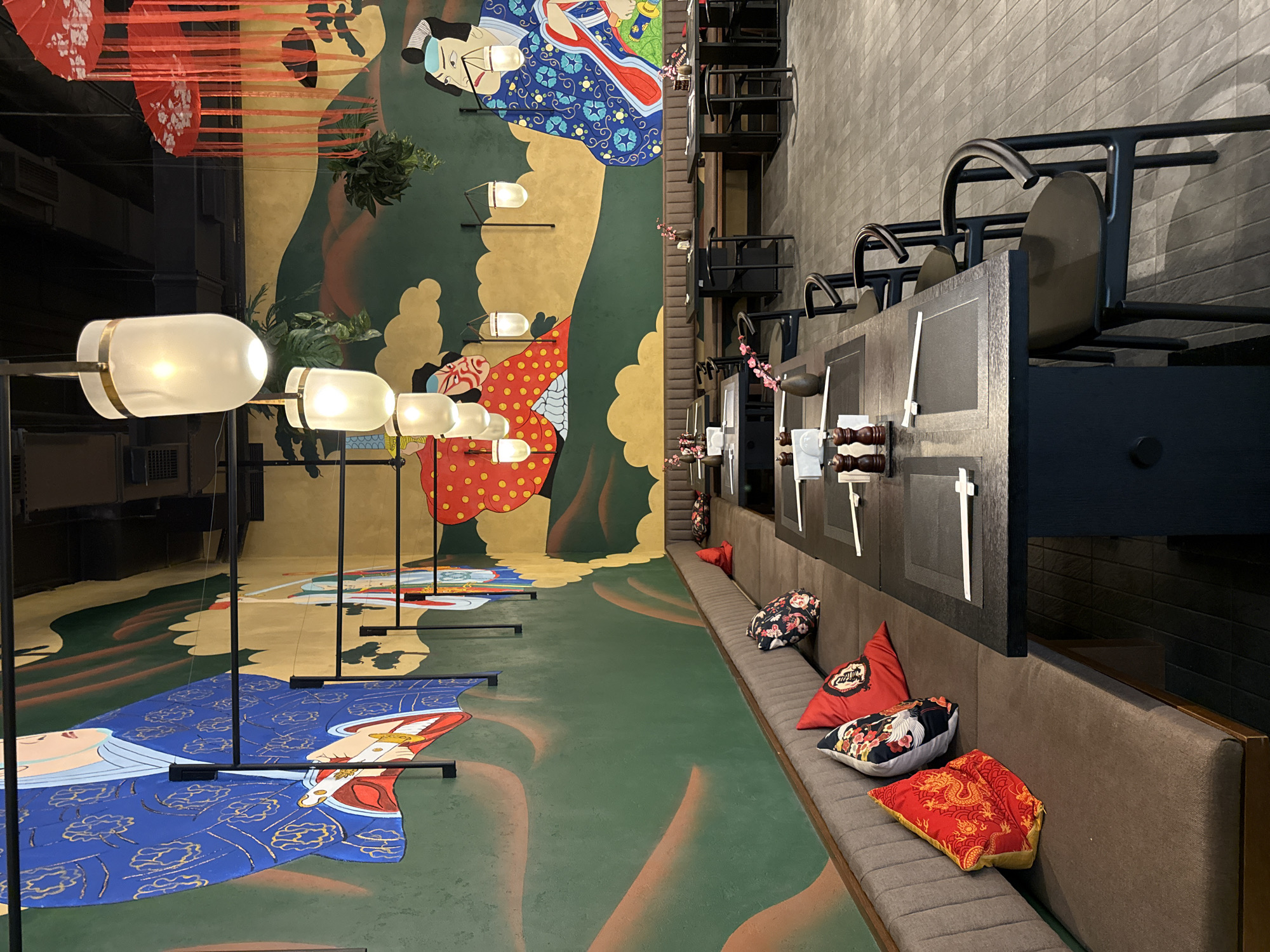 This image captures the interior of an elaborately decorated venue, likely a restaurant or bar, with a strong Asian aesthetic. The scene is dominated by a large, colorful mural that spans from the floor up one wall, depicting stylized figures in traditional Japanese or Chinese attire with flowing robes and intricate patterns, such as a blue kimono with gold cloud designs and a red garment with white polka dots. The floor itself is painted in shades of green and brown, blending seamlessly with the mural's landscape elements.
Elongated, frosted glass light fixtures, mounted on sleek black stands, illuminate the mural from the foreground. Red paper umbrellas, adorned with white floral motifs, are suspended from the ceiling in the upper left, adding to the cultural ambiance.
On the right side of the room, a long, plush grey upholstered booth runs along a textured grey wall, providing extensive seating. Decorative throw pillows in vibrant reds, blacks, and whites, featuring Asian-inspired patterns like dragons, cranes, and floral designs, are scattered across the bench. Dark-colored, possibly wooden, partitions separate individual seating sections along the booth, with black chairs or stools visible behind them. Small sprigs of pink cherry blossoms are placed as delicate accents on these partitions.
The space is empty of people, suggesting a moment before or after service, or a design showcase. The interior lighting indicates it is likely evening or that the artificial lights are the primary source of illumination. There is no visible text. The overall impression is one of immersive cultural decor, creating a unique and visually rich environment.