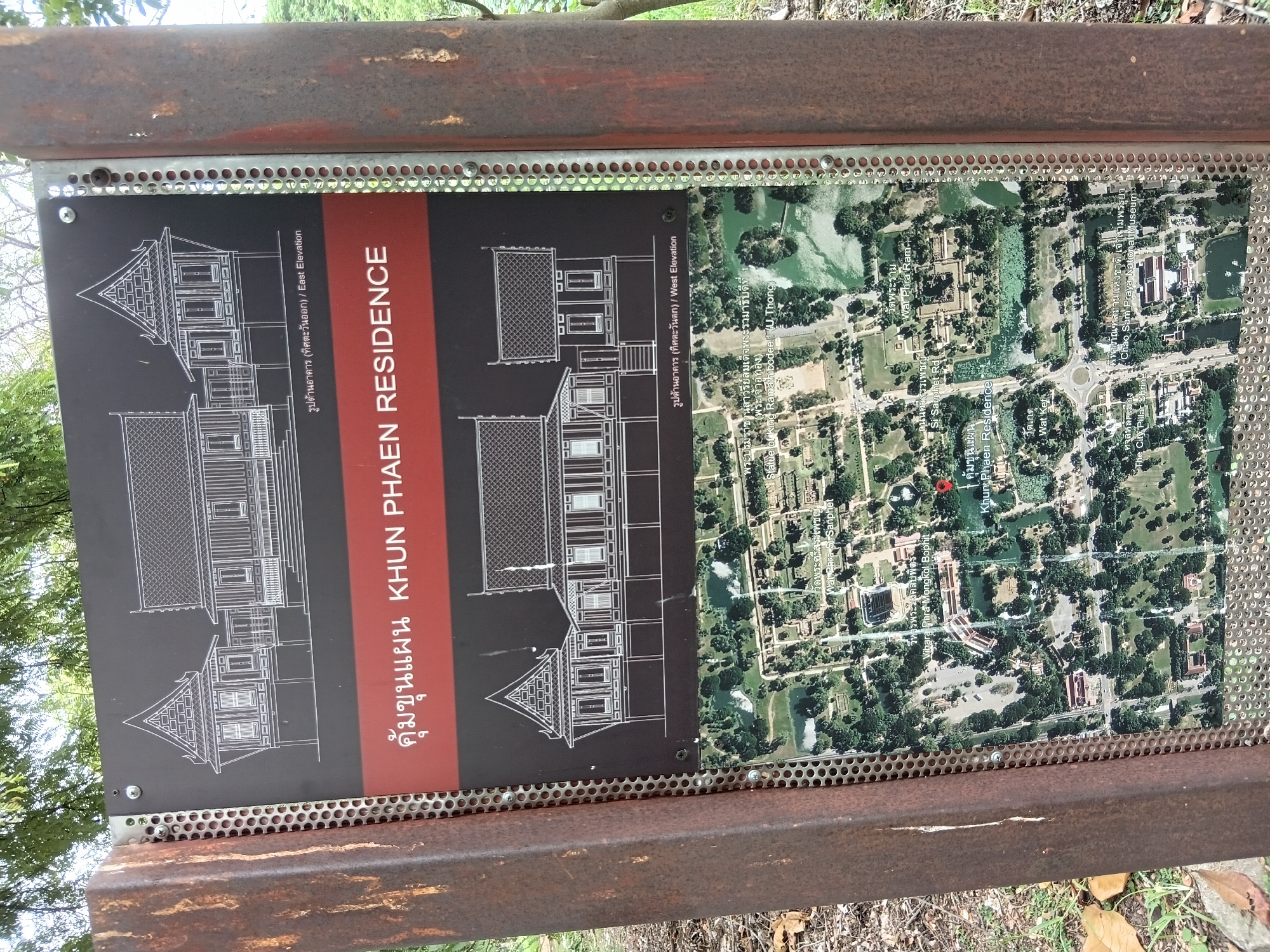 The image shows an informational sign in Phra Nakhon Si Ayutthaya City Municipality, Thailand. The sign has three sections. The top section displays an architectural drawing of a traditional Thai building, labeled in Thai and English as "East Elevation." Below this is a red band with the text "Khun Phaen Residence" in English and Thai. The bottom section of the sign is a satellite map showing the location of the Khun Phaen Residence within a larger area, which includes other landmarks like Wat Phra Si Sanphet, Wat Phra Ram, and the Chao Sam Phraya National Museum. A red marker on the map indicates the precise location of the Khun Phaen Residence. The sign is mounted on a rusty metal post, and the background shows foliage from trees and dry leaves on the ground, suggesting an outdoor setting.