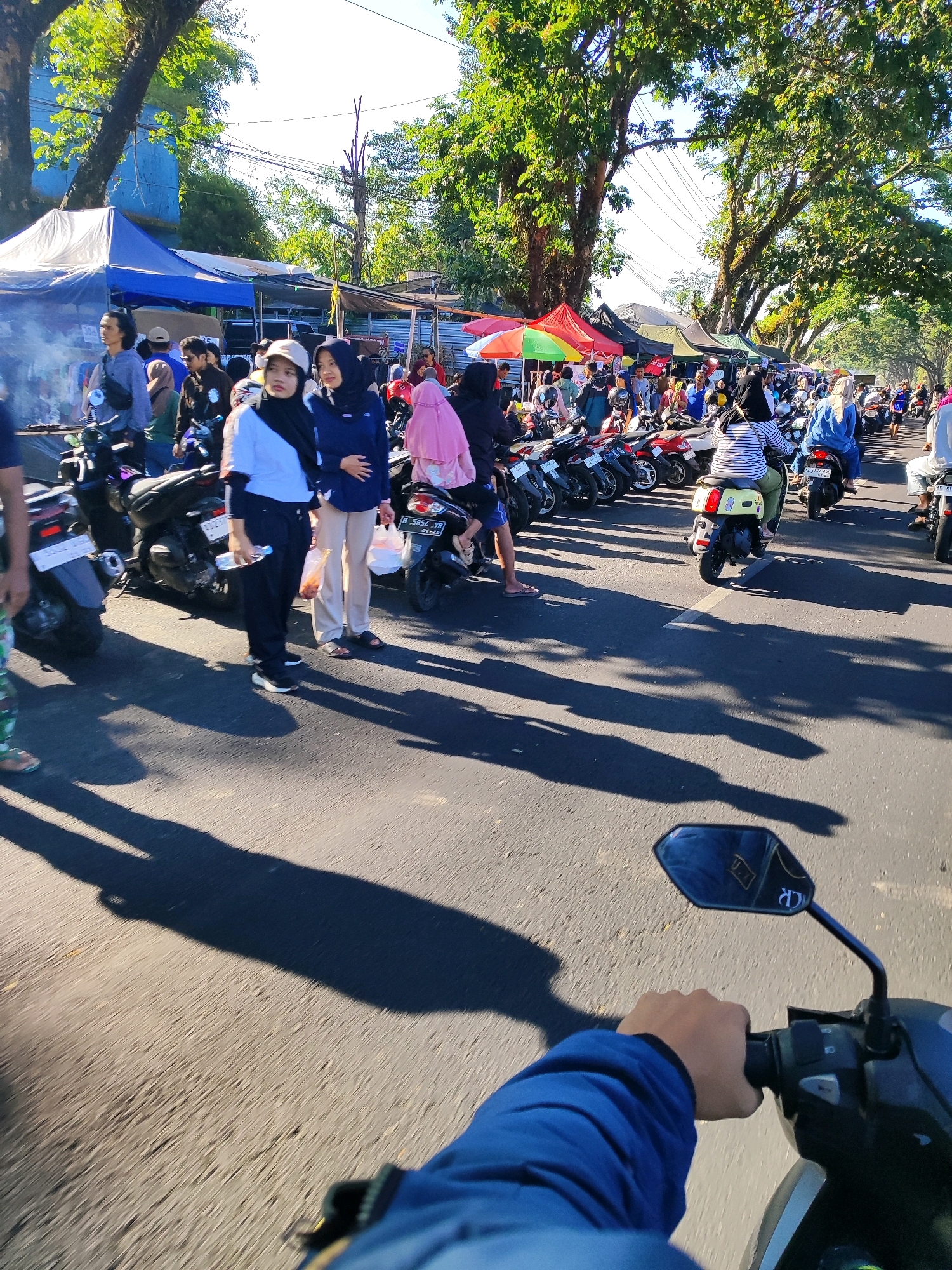 The image depicts a bustling street scene in Salatiga, Indonesia, possibly during a market or festival. The foreground shows a person's hand on the handlebars of a motorcycle, indicating a first-person perspective of someone riding through the area. The road is lined with numerous parked motorcycles and a row of market stalls with blue and red awnings, selling various goods. People are seen walking around, browsing the stalls, and riding their motorcycles. Many individuals are wearing headscarves, suggesting a Muslim-majority population. The environment is characterized by lush green trees lining the street, casting long shadows on the asphalt under a bright, sunny sky. The overall atmosphere is lively and crowded, indicative of a vibrant community gathering.